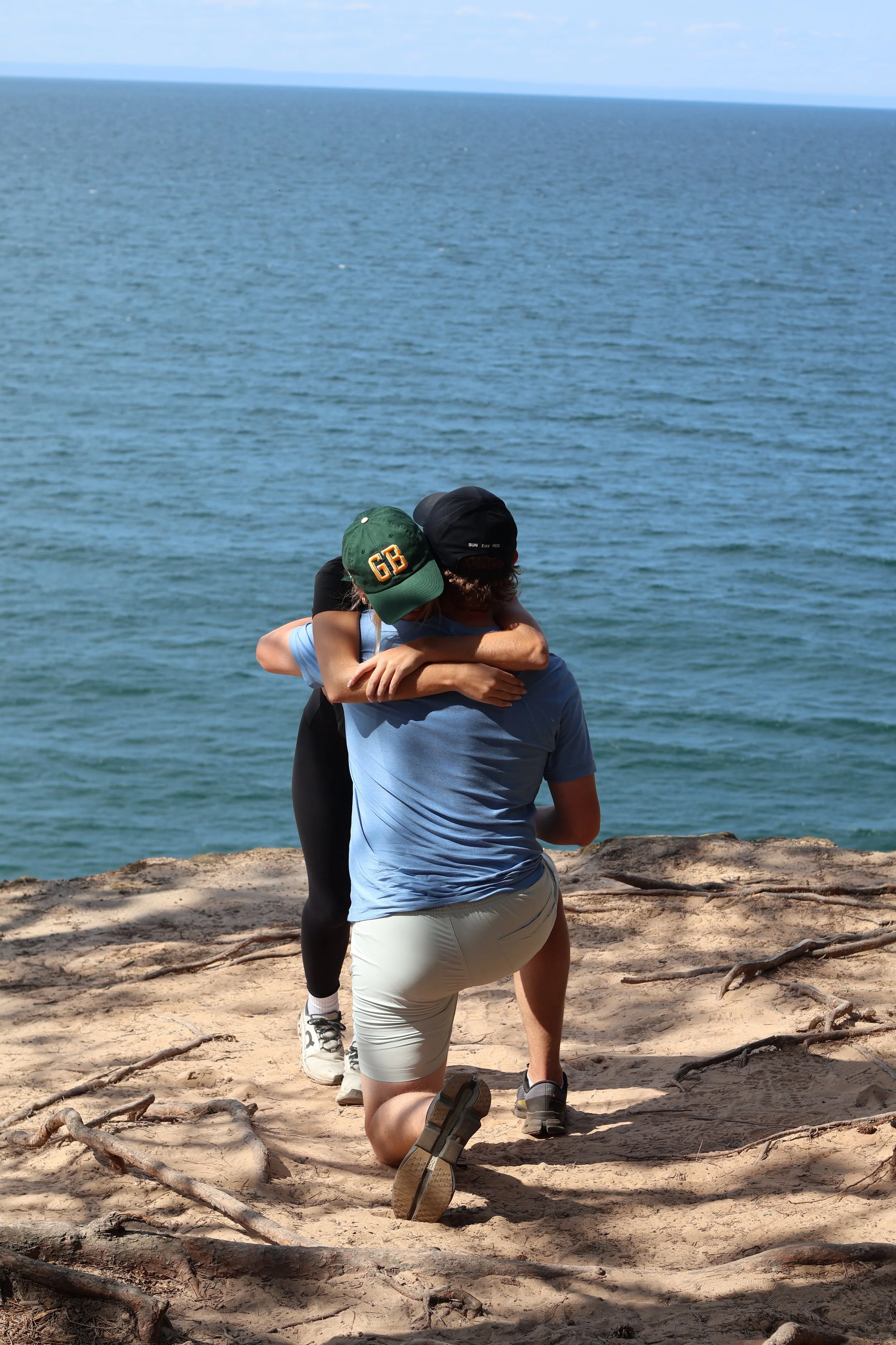 Two people, one in black and the other in light blue, hugging on a sandy beach with driftwood, ocean, and sky in the background.