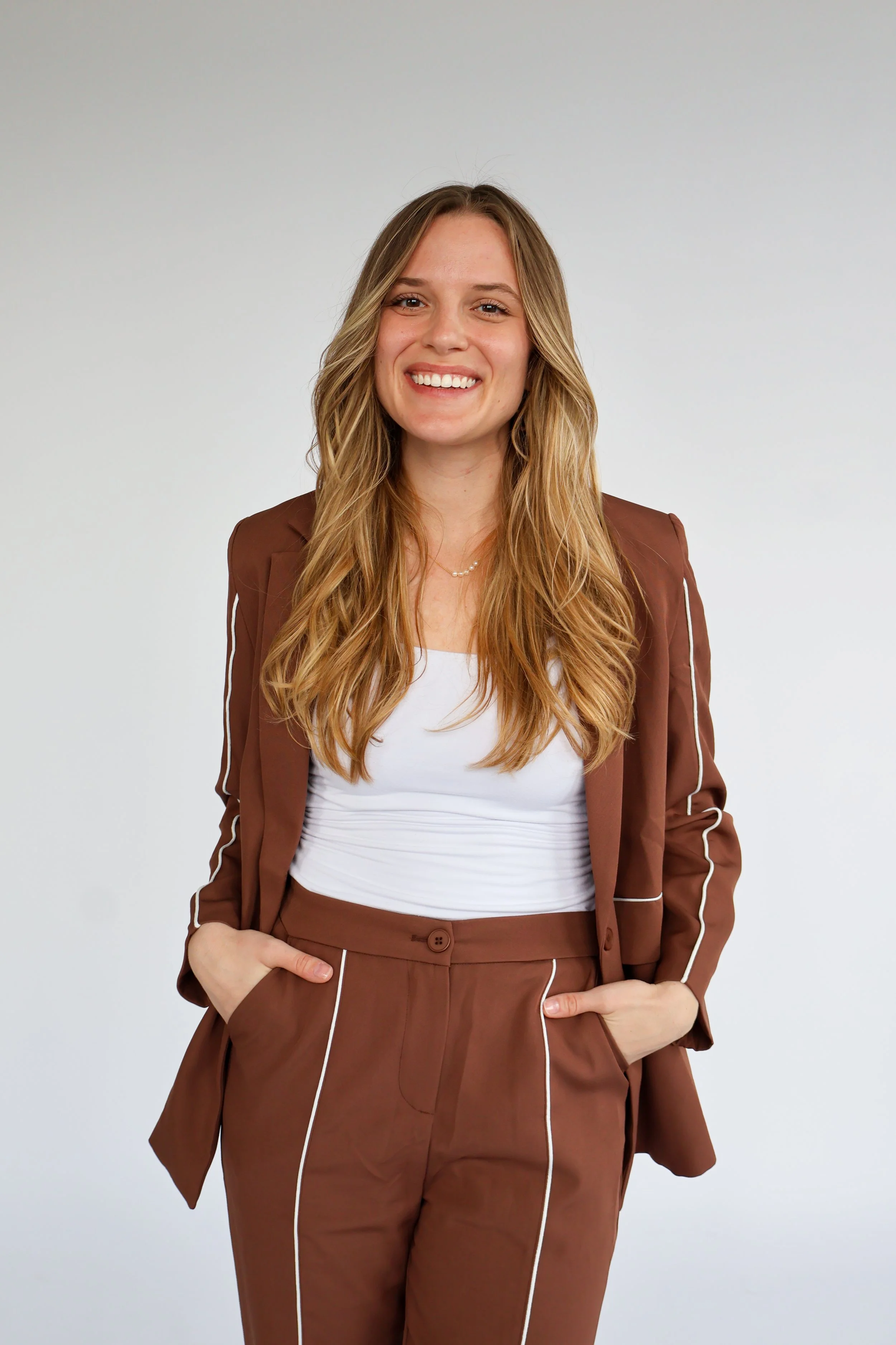 A young woman with long wavy blonde hair, smiling, wearing a brown suit with white piping, white top, and standing against a plain light grey background.