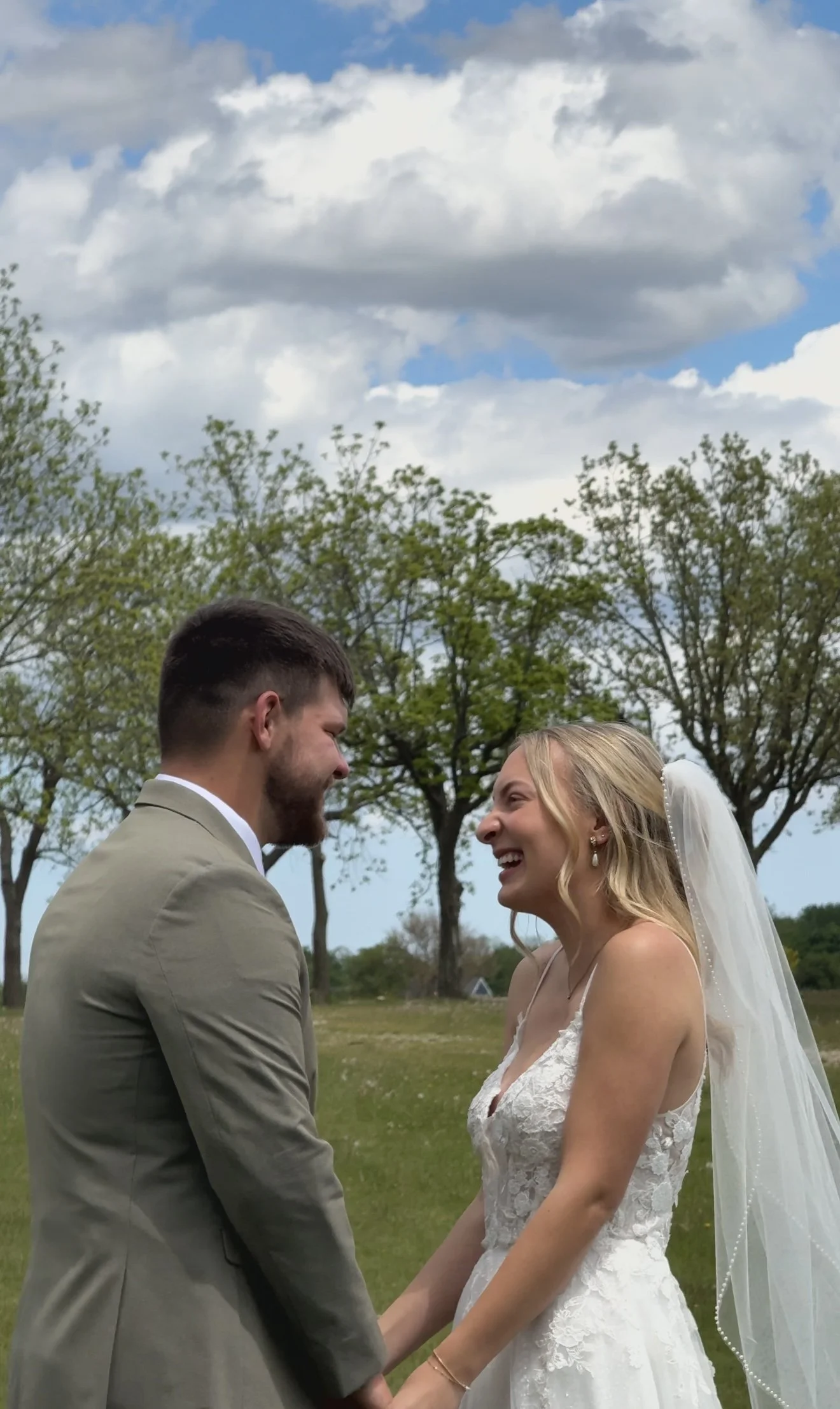 A bride and groom stand outdoors on a grassy field, holding hands and smiling at each other, with trees and a partly cloudy sky in the background.