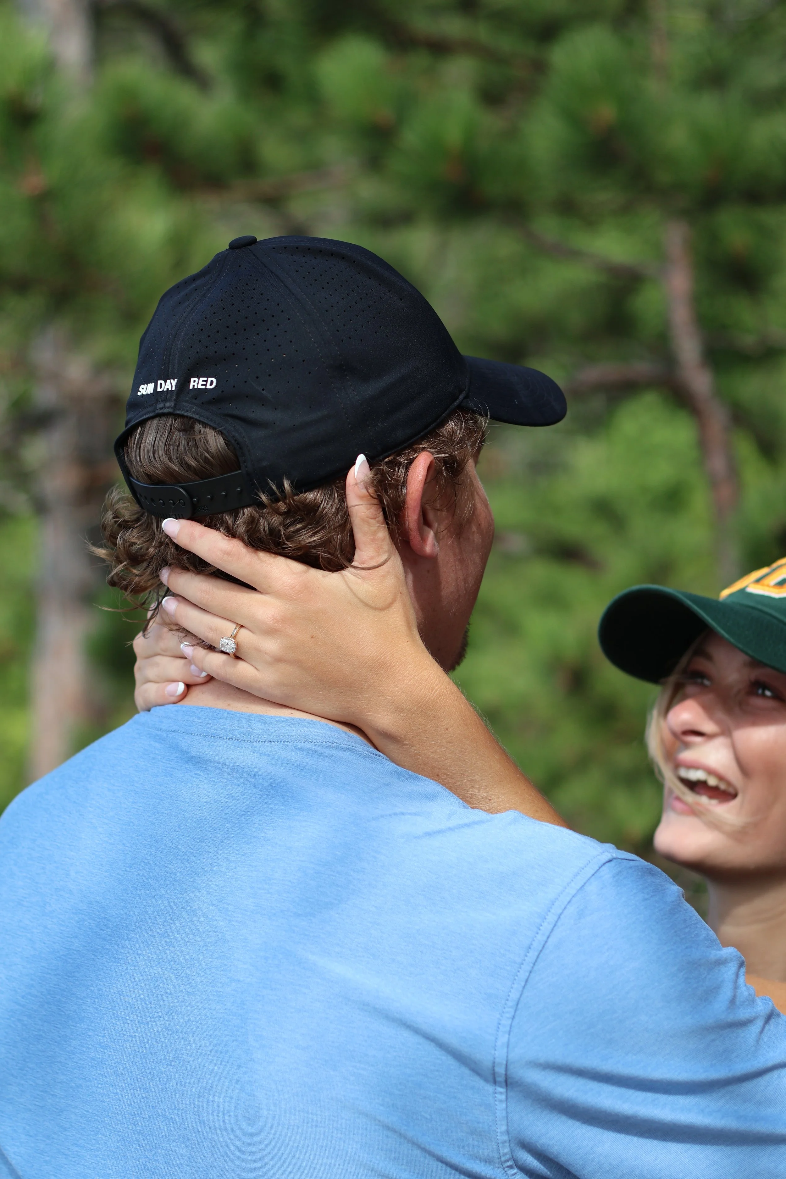 A man and a woman outdoors with trees in the background. The man is wearing a black cap and a light blue shirt, and the woman has long blonde hair and is smiling. The woman is touching the man's neck and face.
