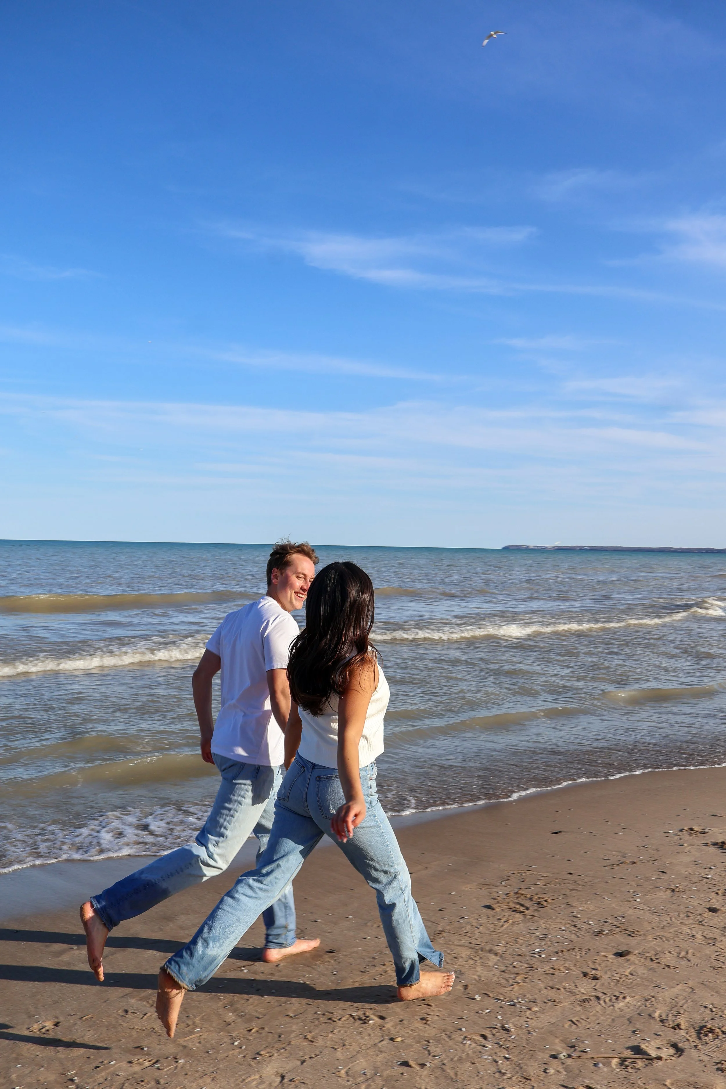Three people, two women and one man, are walking barefoot along the sandy beach near the ocean, with waves gently lapping at their feet, under a bright blue sky with a few wispy clouds and a seagull flying overhead.