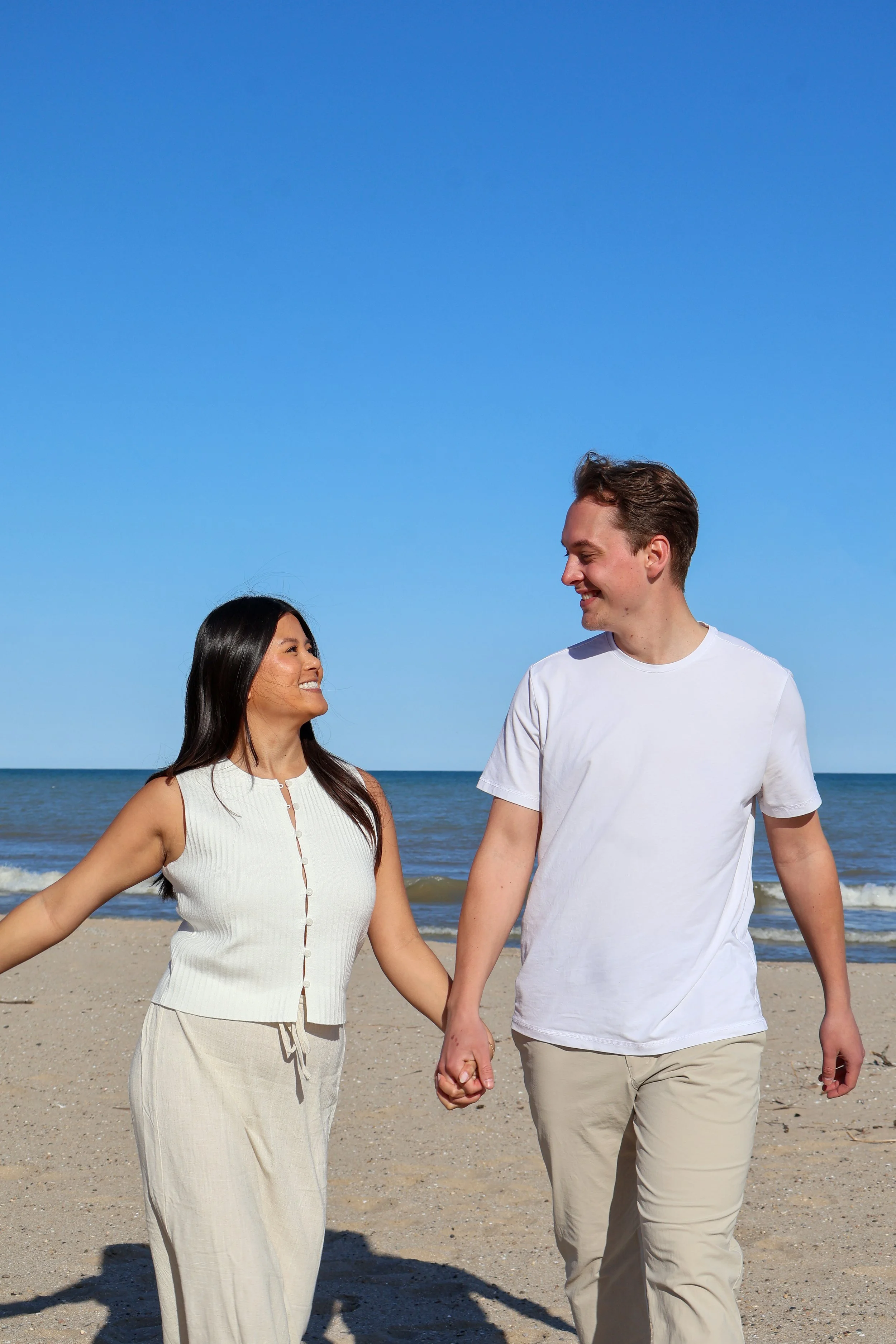 A smiling couple walking on the beach holding hands, with the ocean and blue sky in the background.
