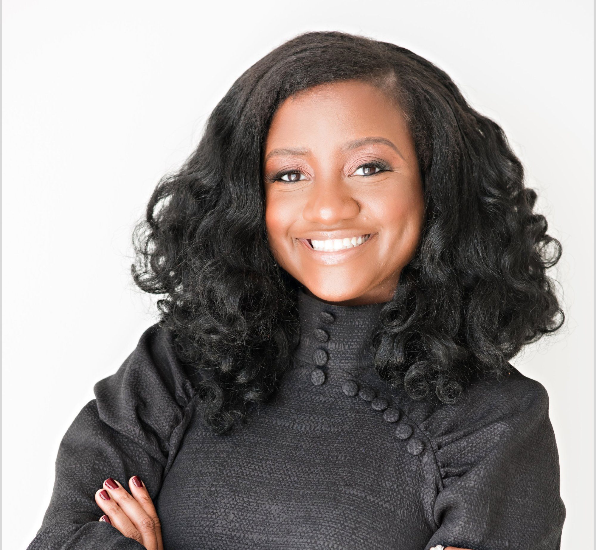 Close-up of an African American woman with curly black hair, smiling, wearing a black textured sweater, with her arms crossed.