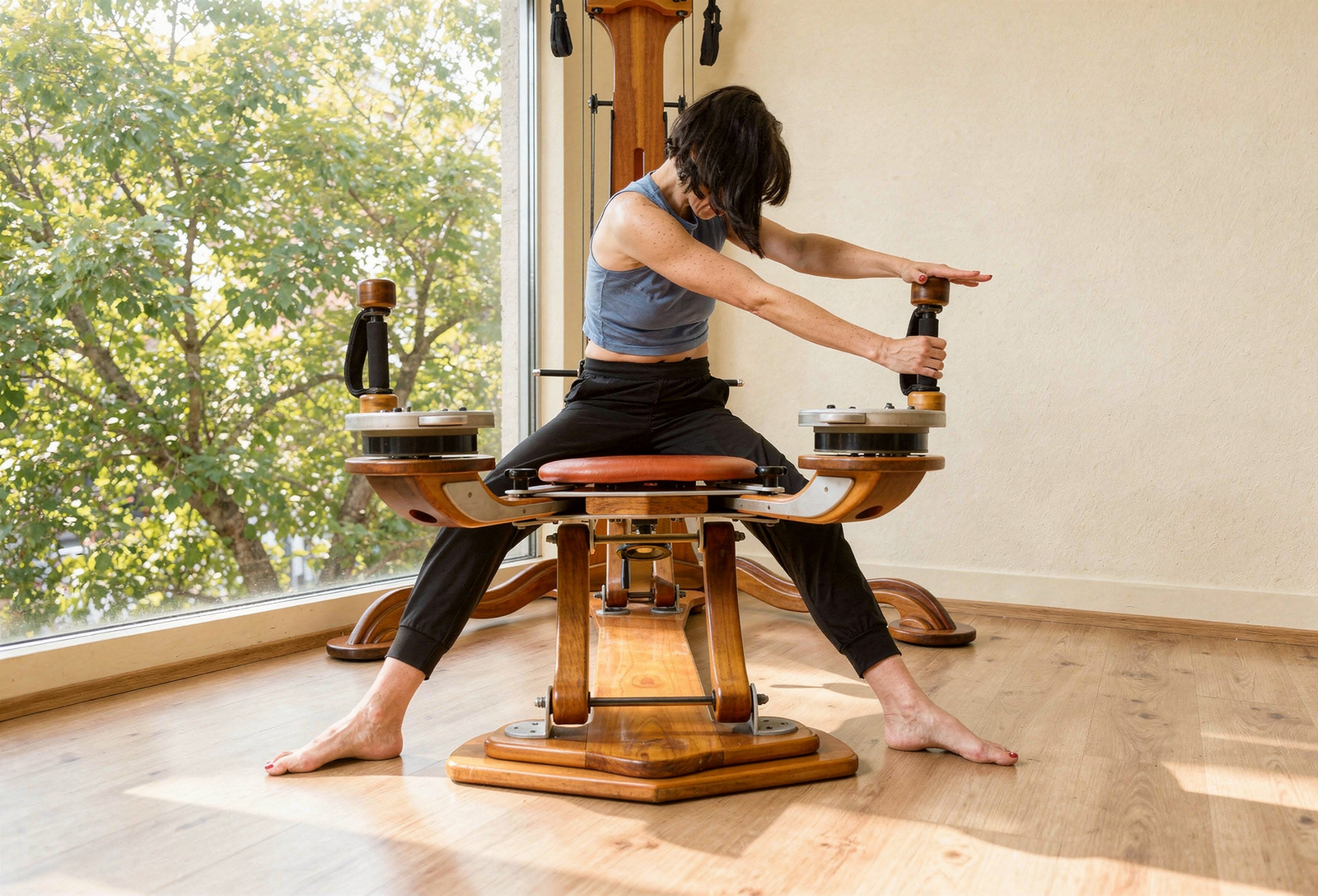 A woman with short black hair, wearing a blue sleeveless top and black pants, is sitting on a wooden massage chair with her arms extended forward, stretching. She is barefoot, and the room has wood flooring and large window with green trees outside, allowing natural light to enter.