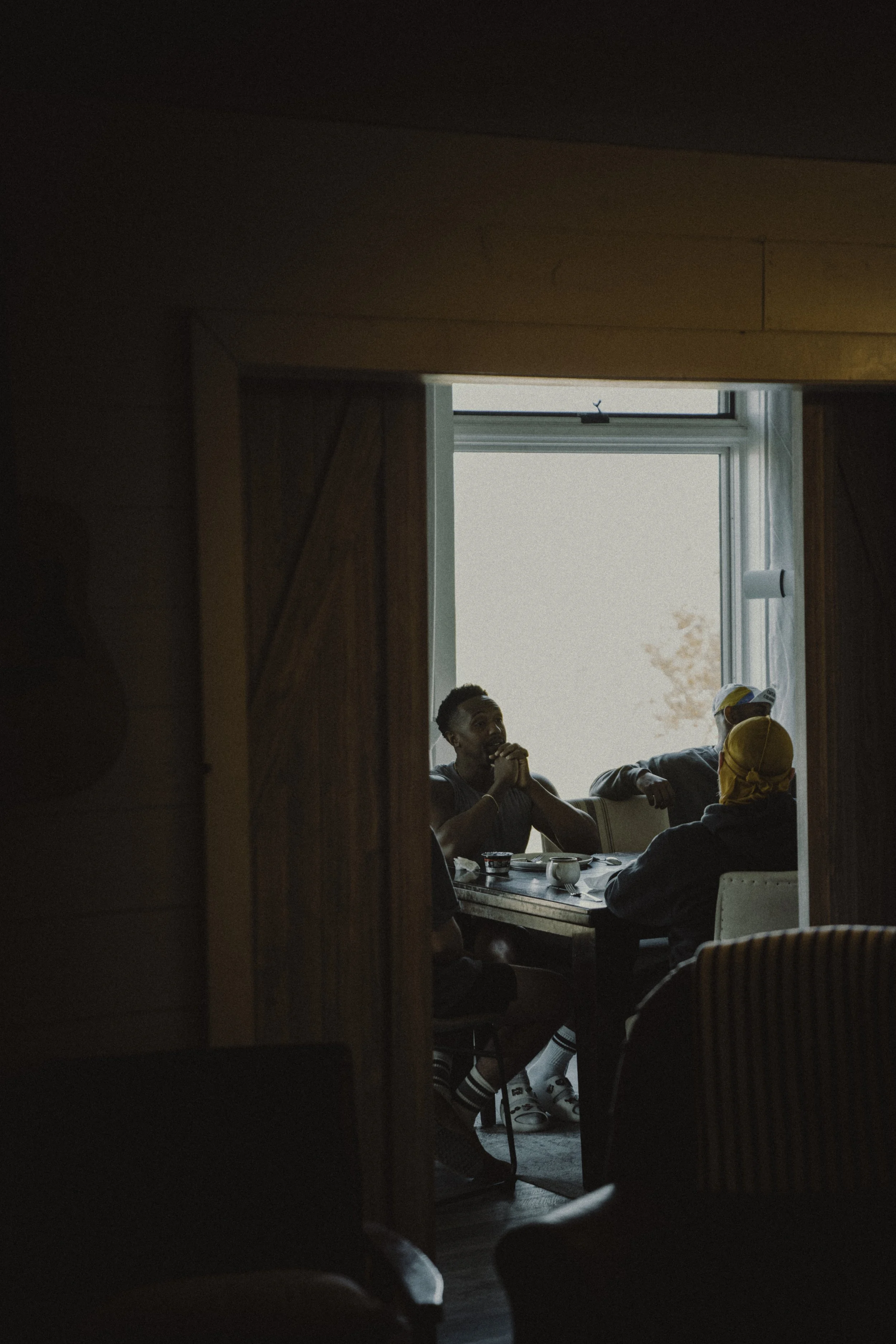 People sitting at a table in a sunlit room, viewed through a doorway with wooden framing, having a discussion and enjoying a moment together. This is ministry--table ministry. A special time of communion and faith-building in a lonely world.