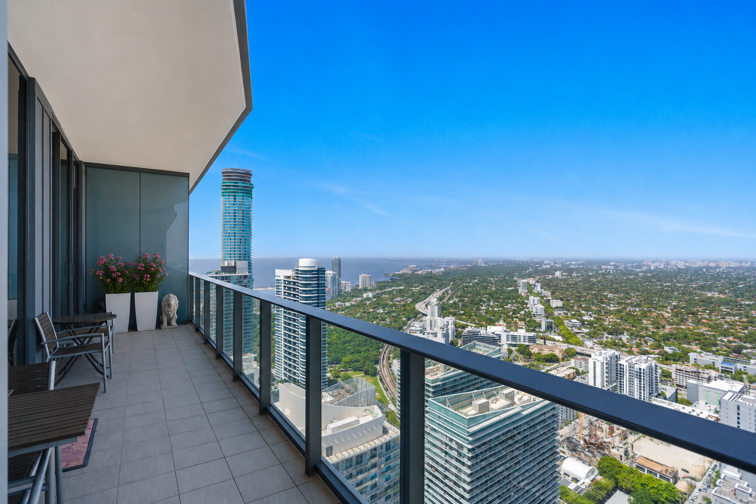 City skyline view from a high-rise balcony, with tall buildings, a clear blue sky, and a body of water in the distance.