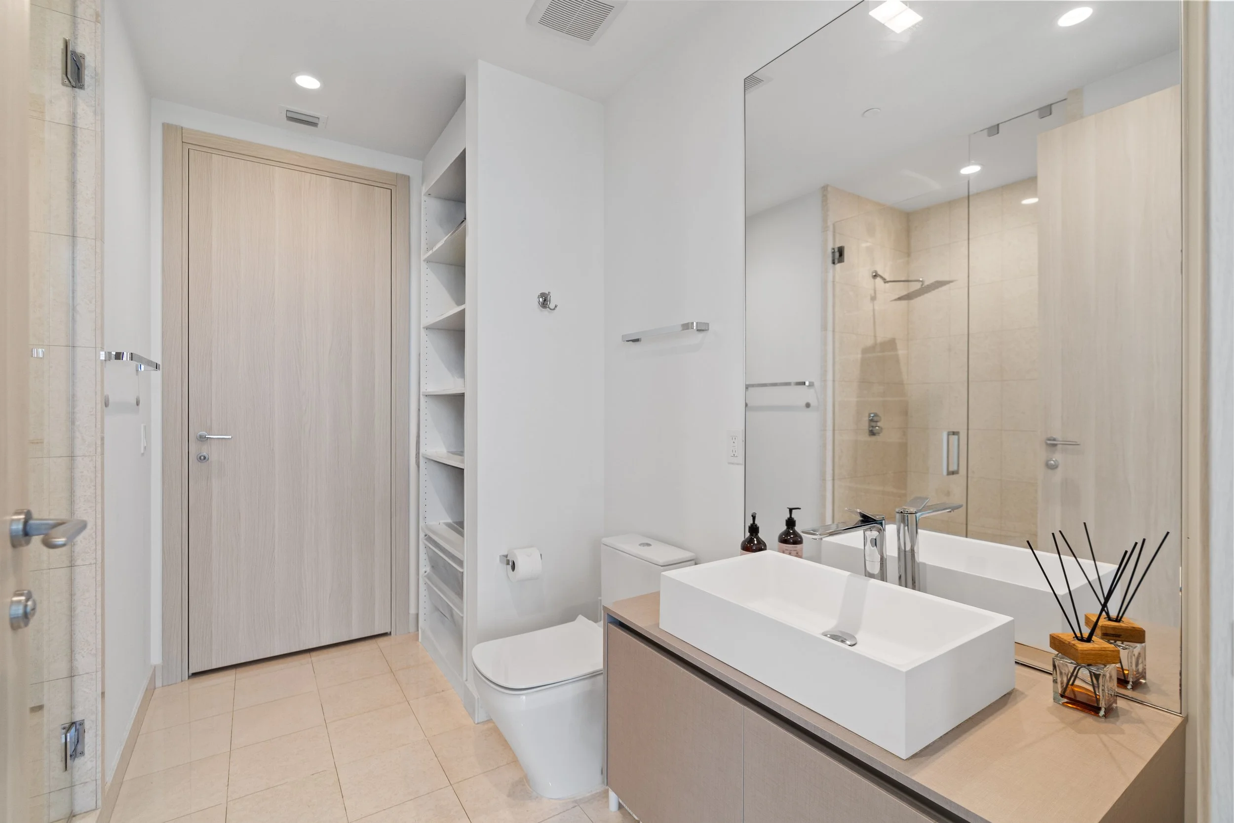 Modern bathroom with a large mirror, white sink, beige tiles, and glass shower enclosure.