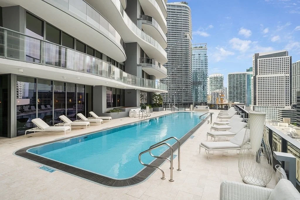 Luxury rooftop swimming pool with lounge chairs, surrounded by modern high-rise buildings under a blue sky.