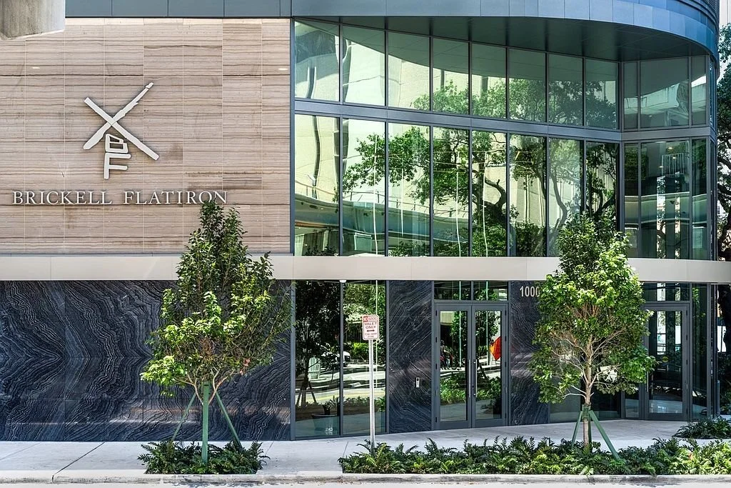 Modern building with glass exterior and trees in front, signage reads 'Brickell Flatiron'