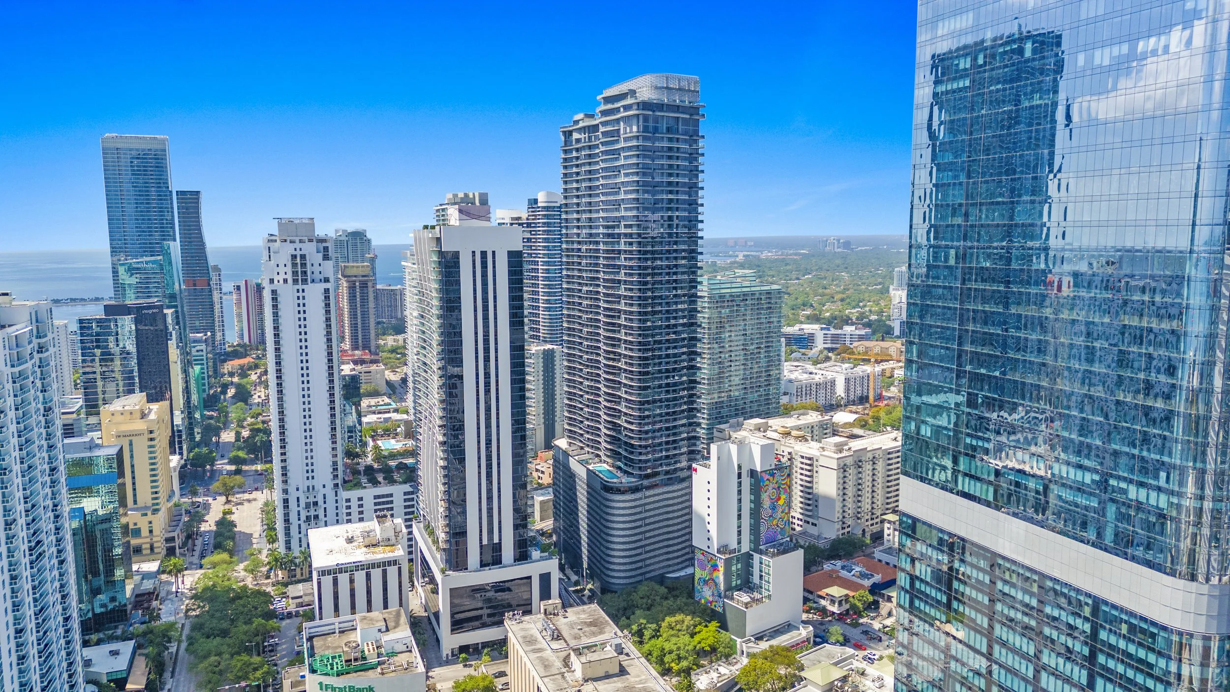 View of a city skyline with tall modern glass skyscrapers and buildings, clear blue sky, some green parks, and the ocean in the background.