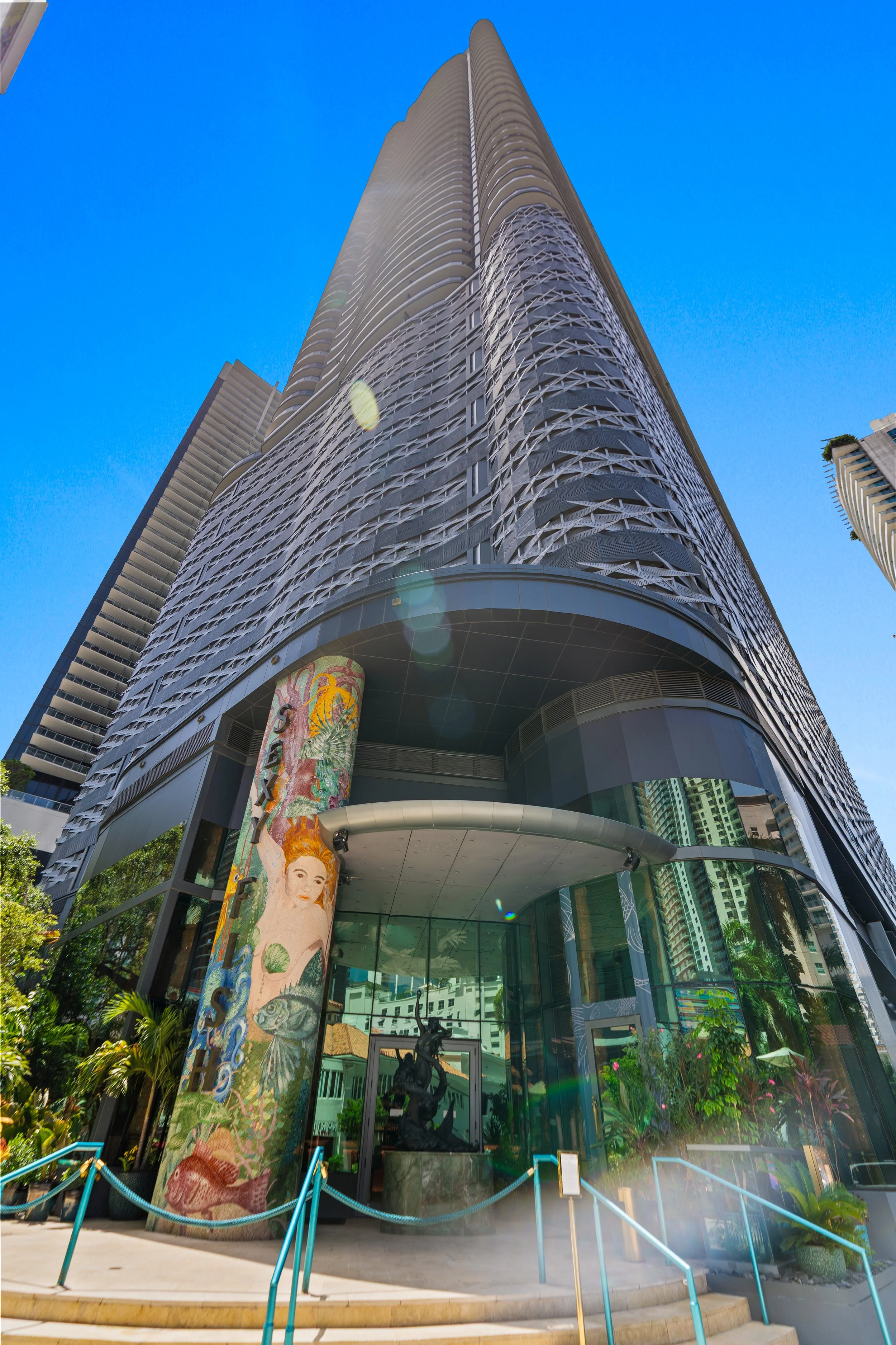 Exterior view of a modern high-rise building with curved architectural design and large glass windows, surrounded by plants and a clear blue sky.