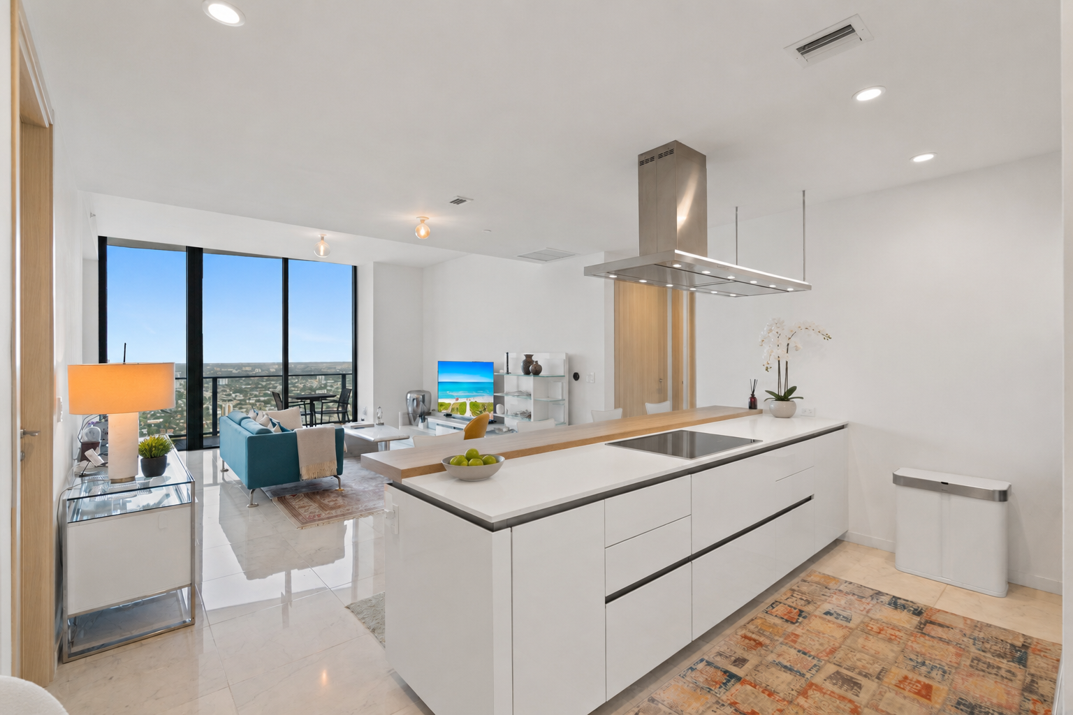 Modern open-concept living space with kitchen island in the foreground, white walls, and large floor-to-ceiling windows overlooking a city skyline.