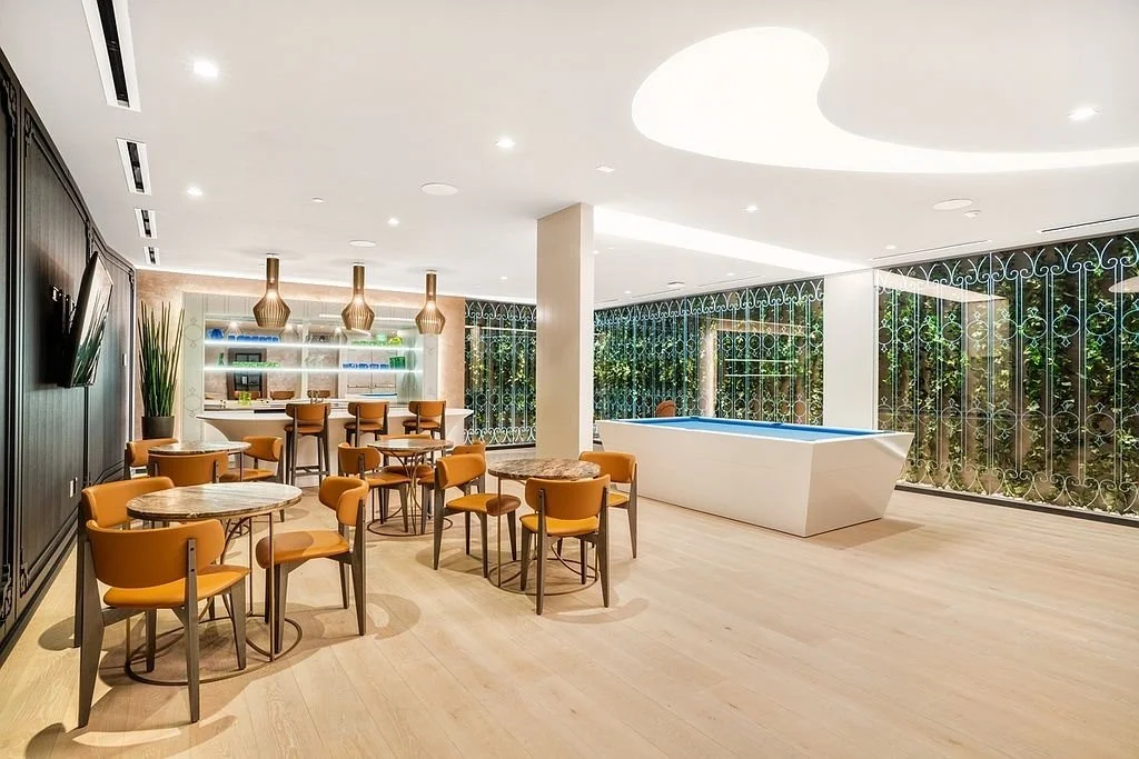 Modern lounge area with round tables and orange chairs, a kitchenette with bar stools, and a large white hot tub, surrounded by large glass windows with decorative metal screens, and bright lighting.