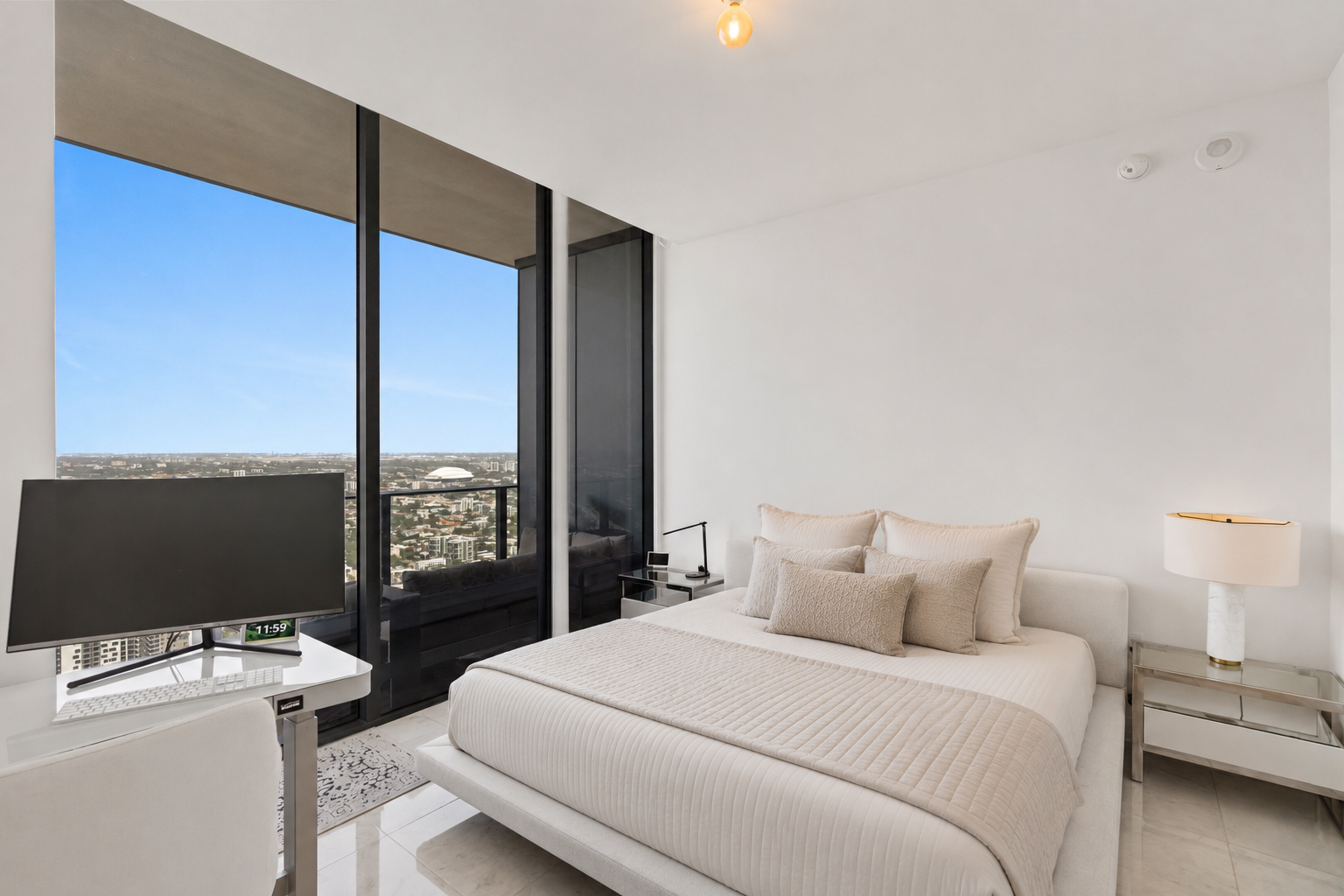 Modern bedroom with white bed, beige pillows, black desk with monitor, and large floor-to-ceiling windows showing a city view.