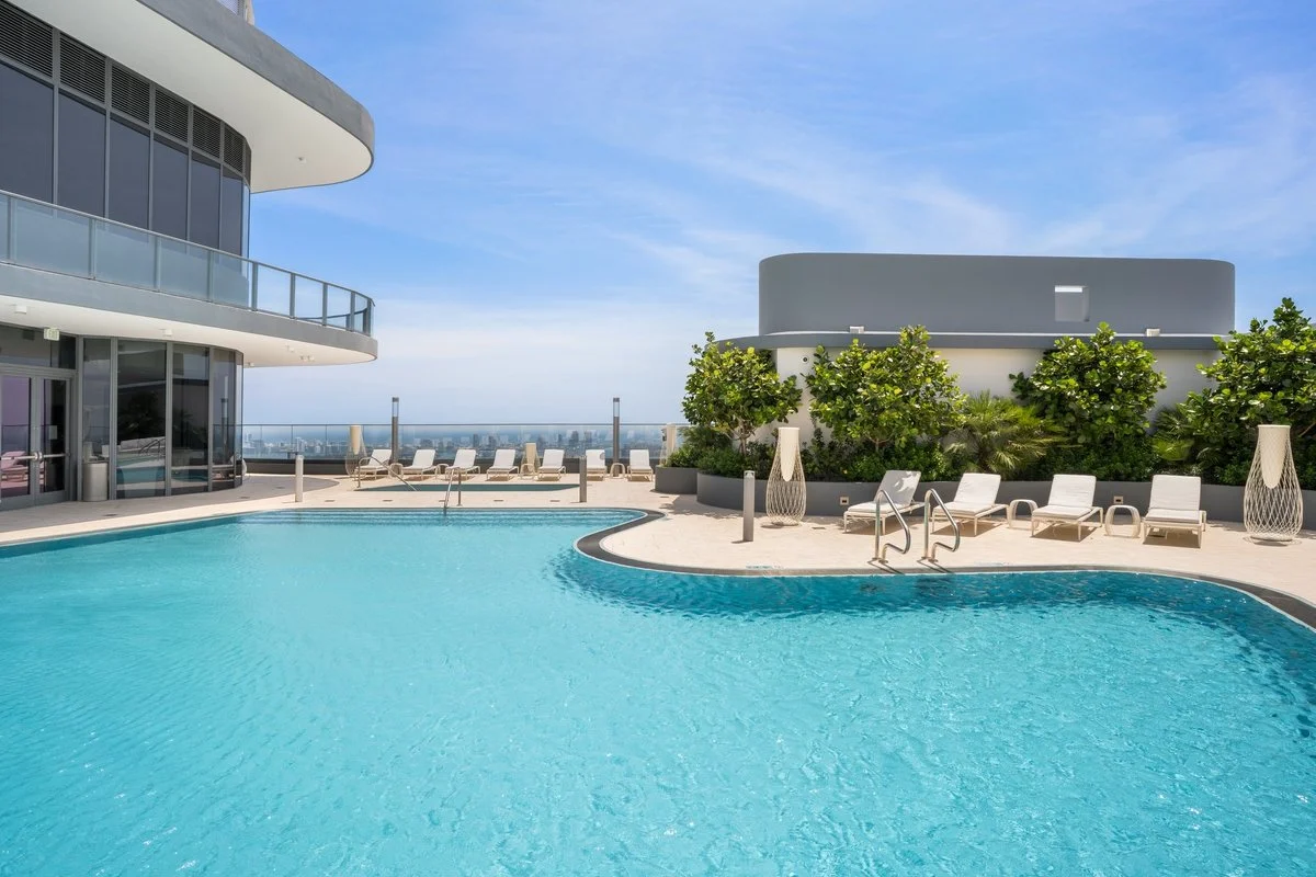 Stylish rooftop swimming pool area with lounge chairs, greenery, and a modern building in the background under a partly cloudy sky.