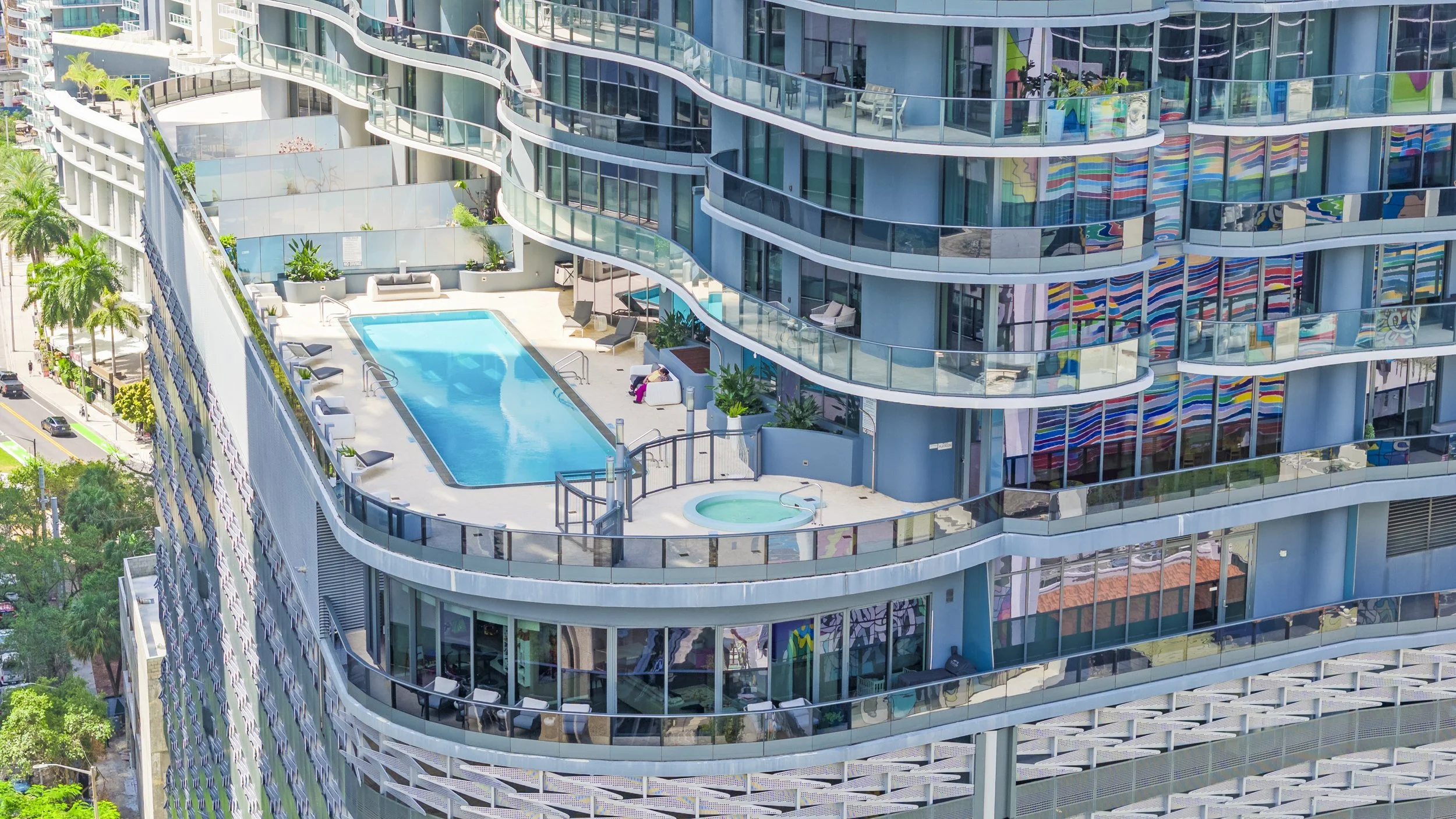 High-angle view of a modern apartment building with curved glass balconies, outdoor swimming pool, and rooftop lounging area.