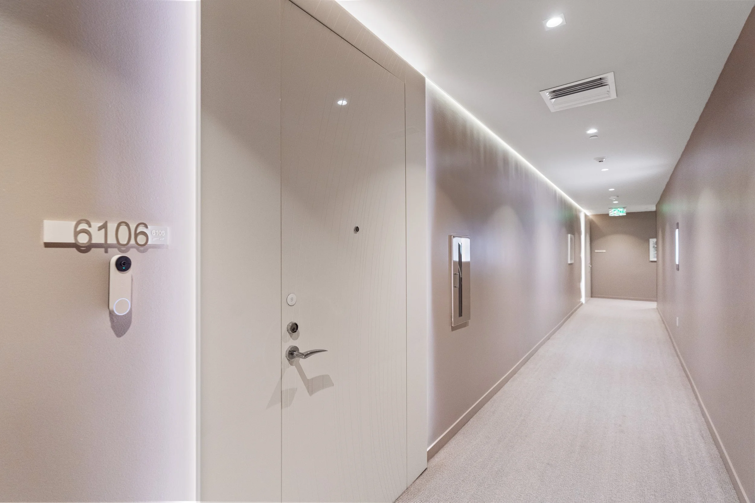 Modern hallway with beige walls, ceiling lights, a door with the number 6106, and a peephole, leading to a distant exit door.