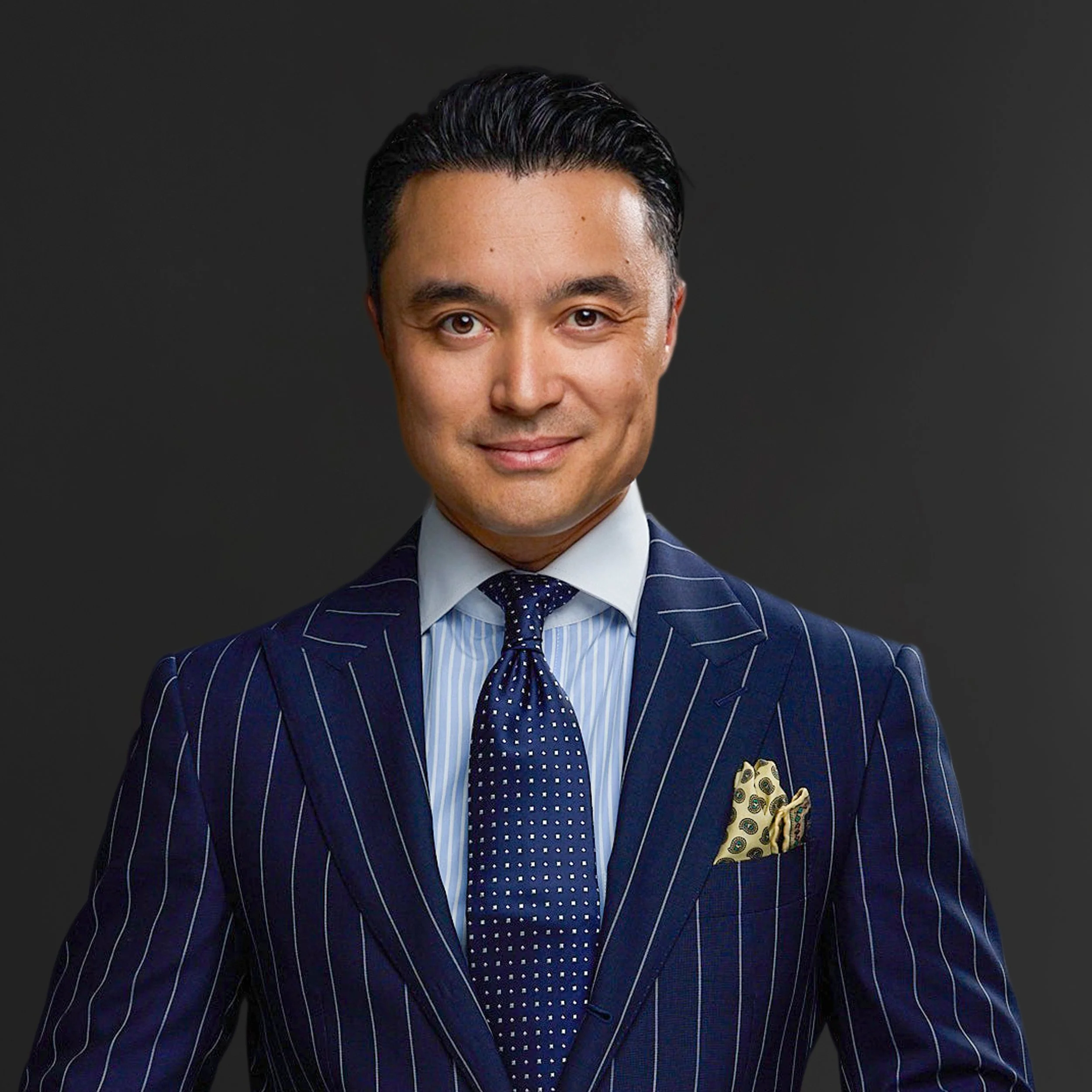 Partial view of a man in a blue pinstripe suit, white shirt, and blue polka-dot tie, with a yellow-patterned pocket square, against a dark background.