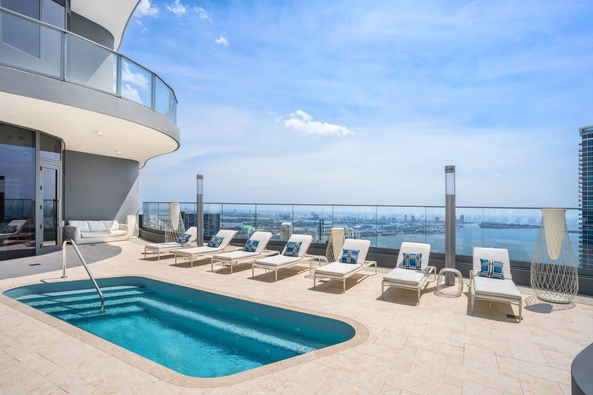 Rooftop pool area with seven white lounge chairs, each with a blue-striped pillow, a small side table, a tall lamp, and a glass barrier overlooking a city skyline with water in the distance, under a partly cloudy sky.