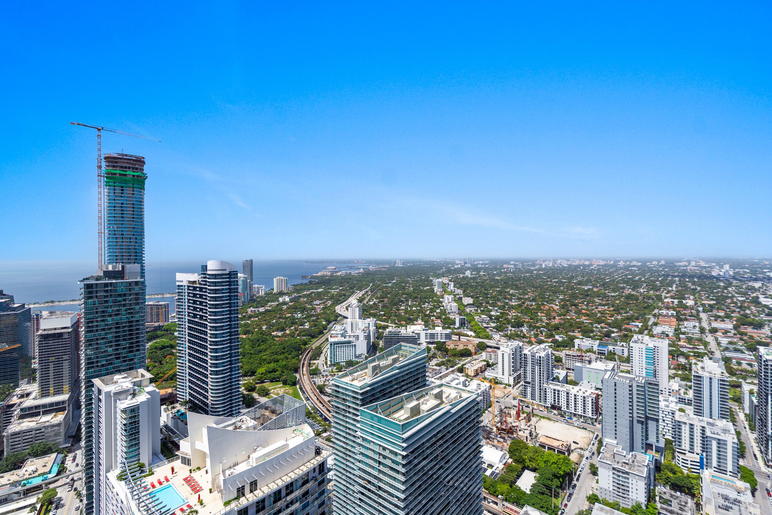 Aerial view of a city with high-rise buildings, a construction site with a tall crane, and a large body of water in the background under a clear blue sky.