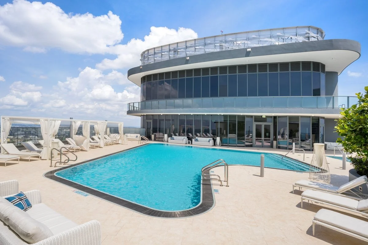 Luxury rooftop pool area with white lounge chairs and cabanas, modern glass building, and blue sky with clouds