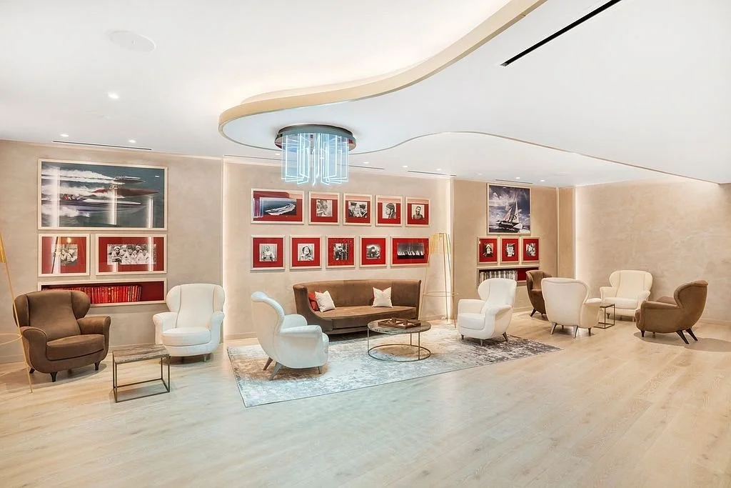 Modern hotel lobby with a mix of brown and white armchairs, a brown sofa, and small side tables. The walls are decorated with framed artwork, including photographs and paintings. There is a unique ceiling light fixture in the center of the room.