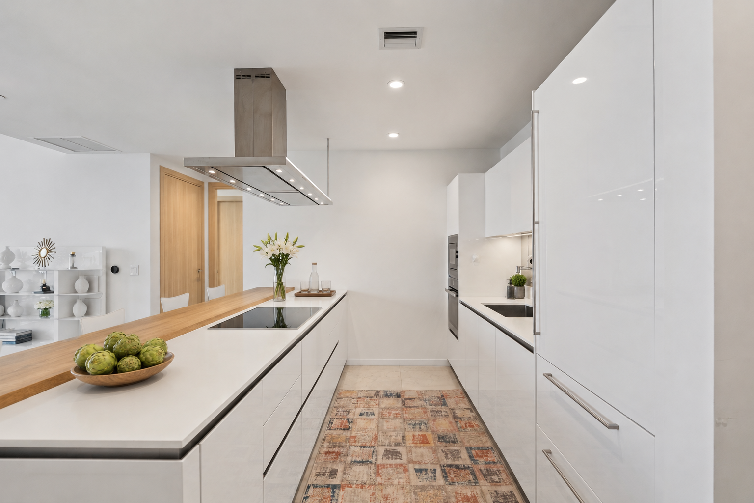 Modern kitchen with white cabinets, wooden accents, built-in appliances, a stovetop on an island, a vase of flowers, artichokes, and a colorful rug.