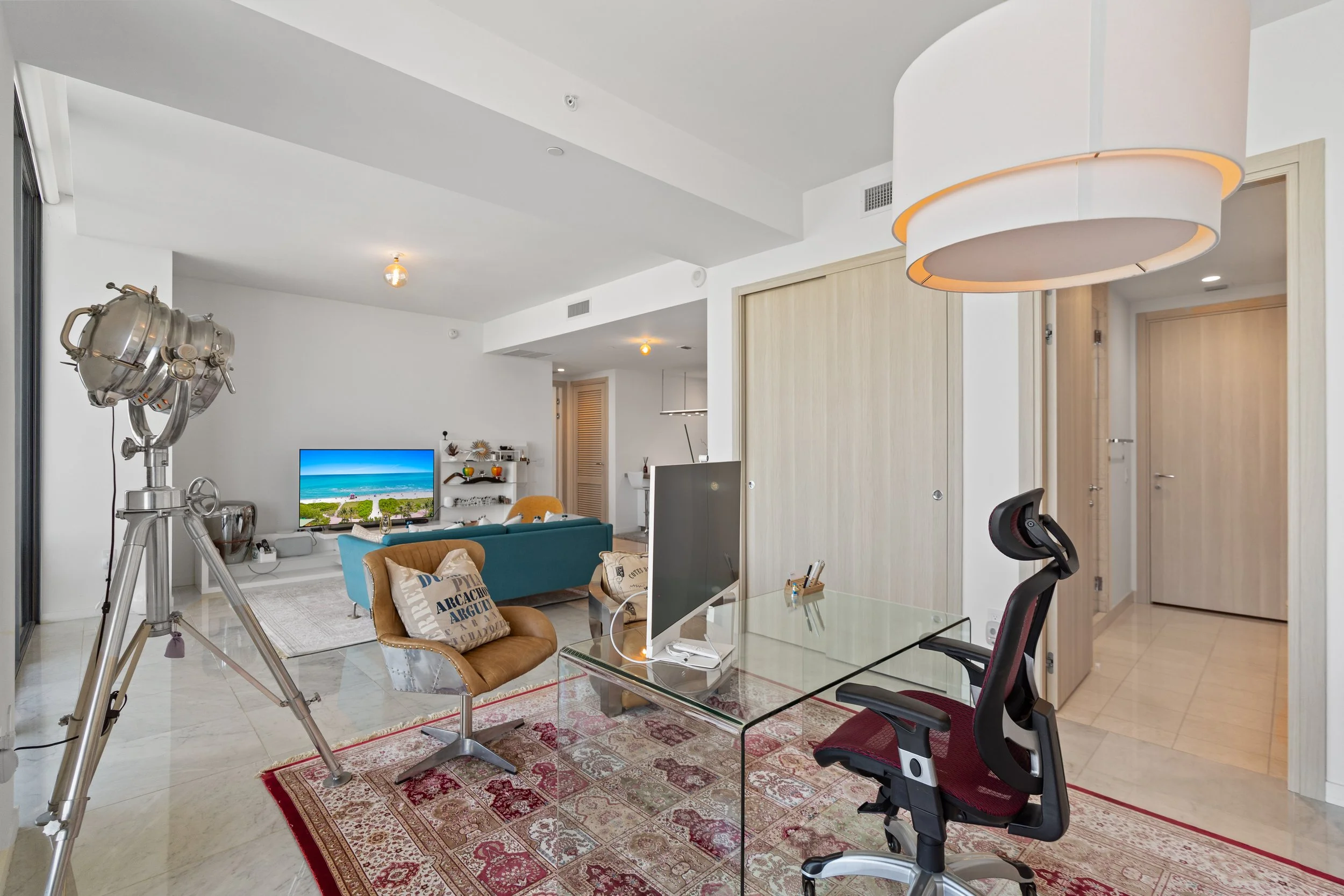 Modern living room with a glass dining table, ergonomic office chair, leather armchair with cushions, blue sofa, flat-screen TV, and decorative items, with a large white hanging light fixture and a vintage spotlight on a tripod.