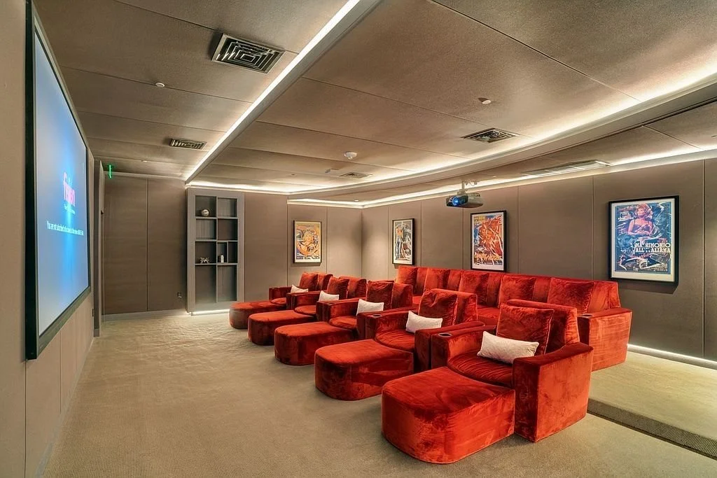 A cozy home theater room with red velvet armchairs arranged in two rows, a large wall-mounted TV on the left, framed movie posters on the wall, and soft ambient lighting.