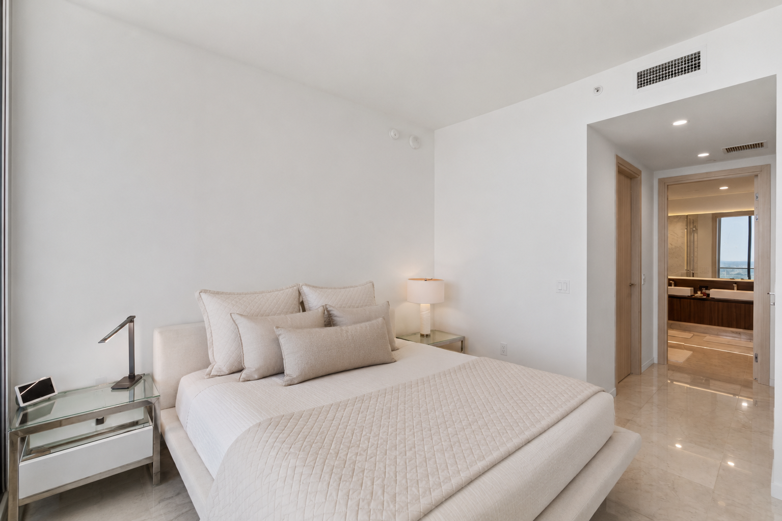 Modern bedroom with white walls, beige bedding, multiple pillows, a bedside lamp, and a glass nightstand with a tablet