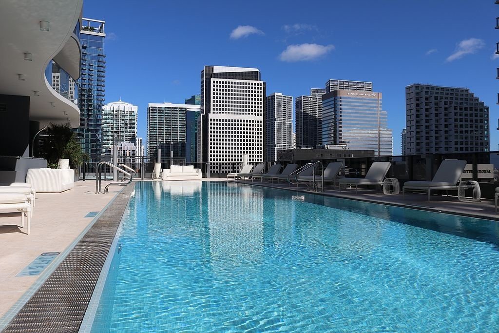 Rooftop swimming pool with lounge chairs and a city skyline background featuring tall buildings under a blue sky.