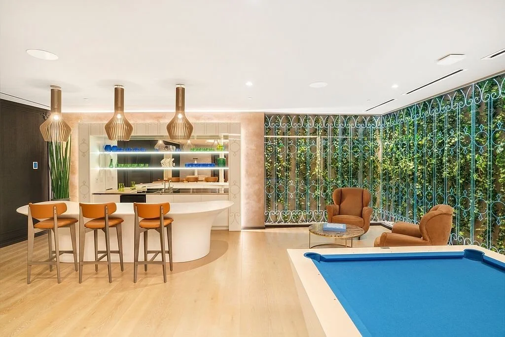 Modern indoor lounge area featuring a bar with four orange chairs, two brown armchairs, a blue pool table, and large windows with decorative metal grilles showing green foliage outside.