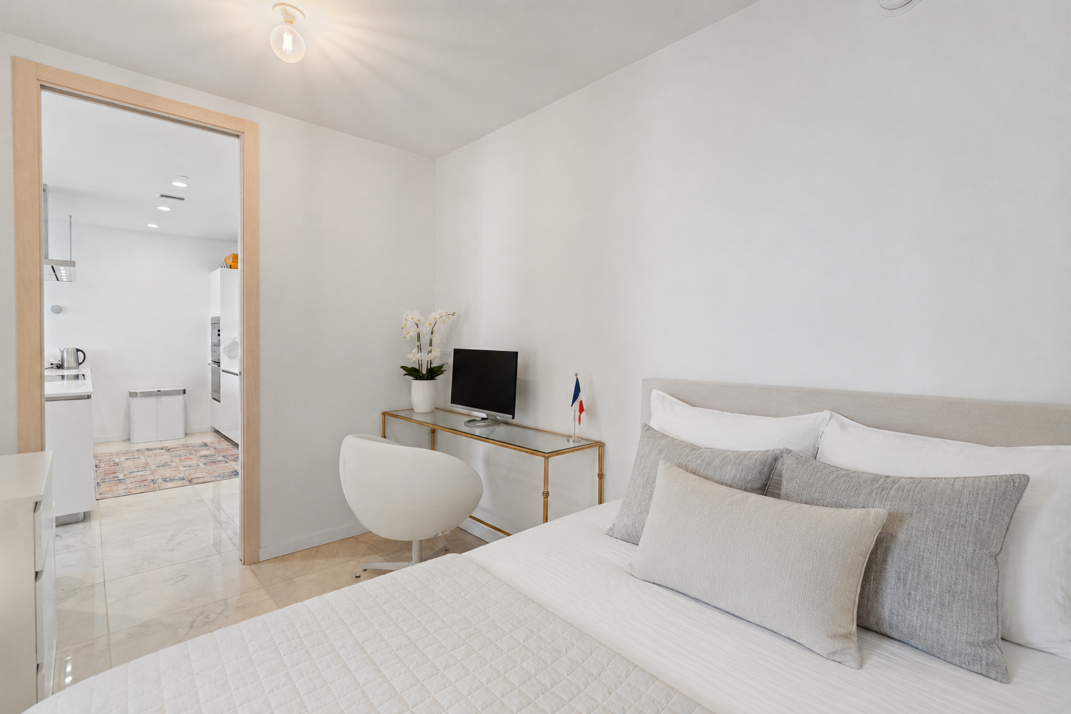 Modern bedroom with white bedding, gray and beige pillows, small desk with a TV, a white chair, and a door leading to a kitchenette with a fridge, microwave, kettle, and a small rug.