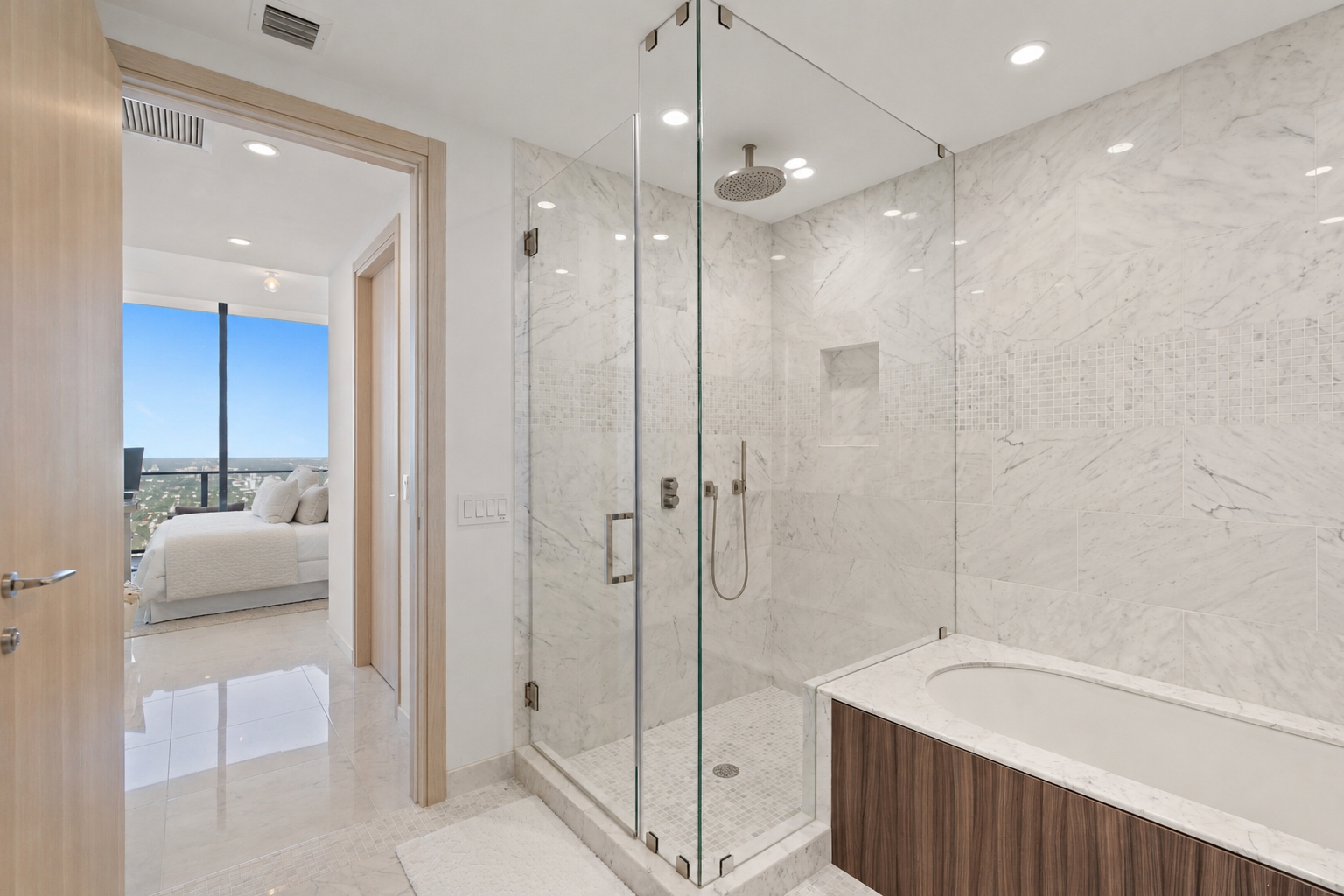 Modern bathroom with marble walls and bathtub, glass-enclosed shower with showerhead and handheld attachment, view into bedroom with large window showing cityscape.
