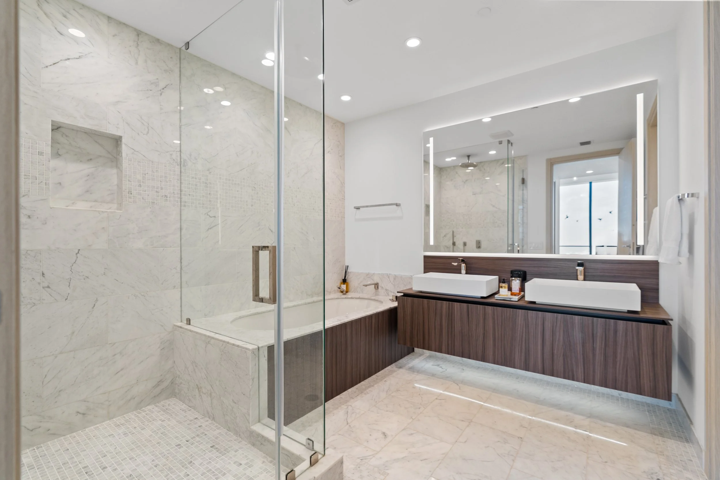 Modern bathroom with a walk-in shower with glass door, marble tiles, and built-in shelf, a double vanity with vessel sinks, large mirror, and contemporary fixtures.