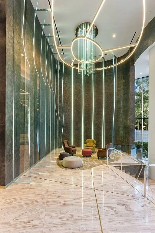 Modern lobby with green marble walls, high ceiling, glass railing, patterned marble flooring, and unique abstract lighting fixture with LED strips and circular pendant lights. Comfortable armchairs and ottomans are arranged around a small rug.