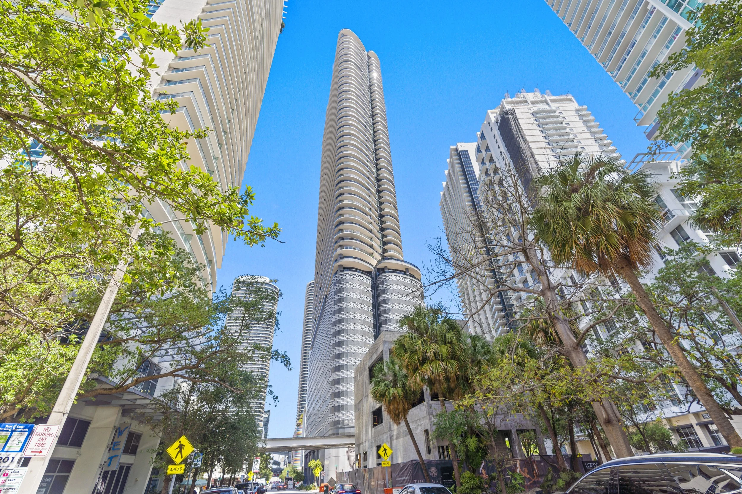 Tall modern skyscrapers with balconies in a city, surrounded by green trees and a clear blue sky.