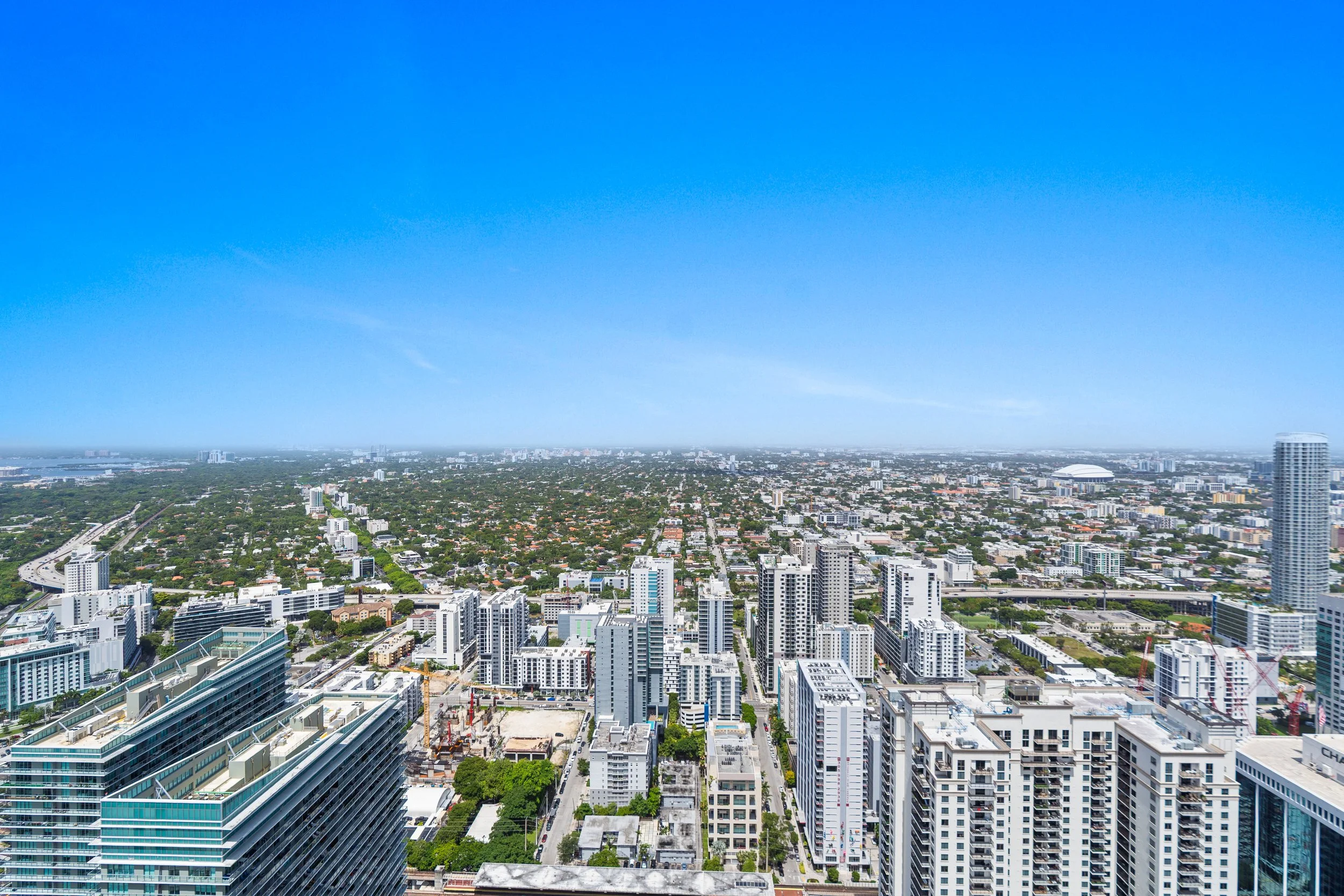 A panoramic view of a city with high-rise buildings, a grid of streets, and a large green park, under a clear blue sky.