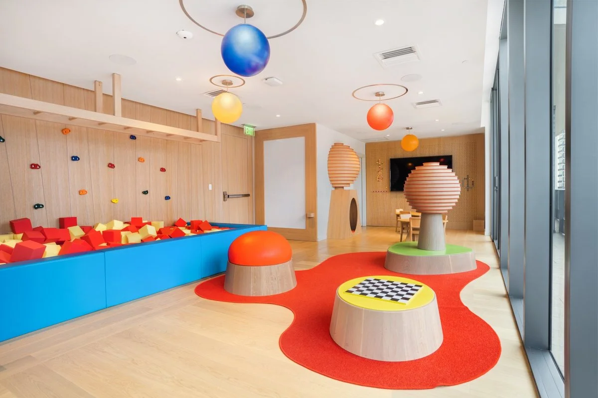 Colorful playroom with foam block pit, climbing wall, large lamps, chess table, and floor seating, bright and welcoming interior with natural light.
