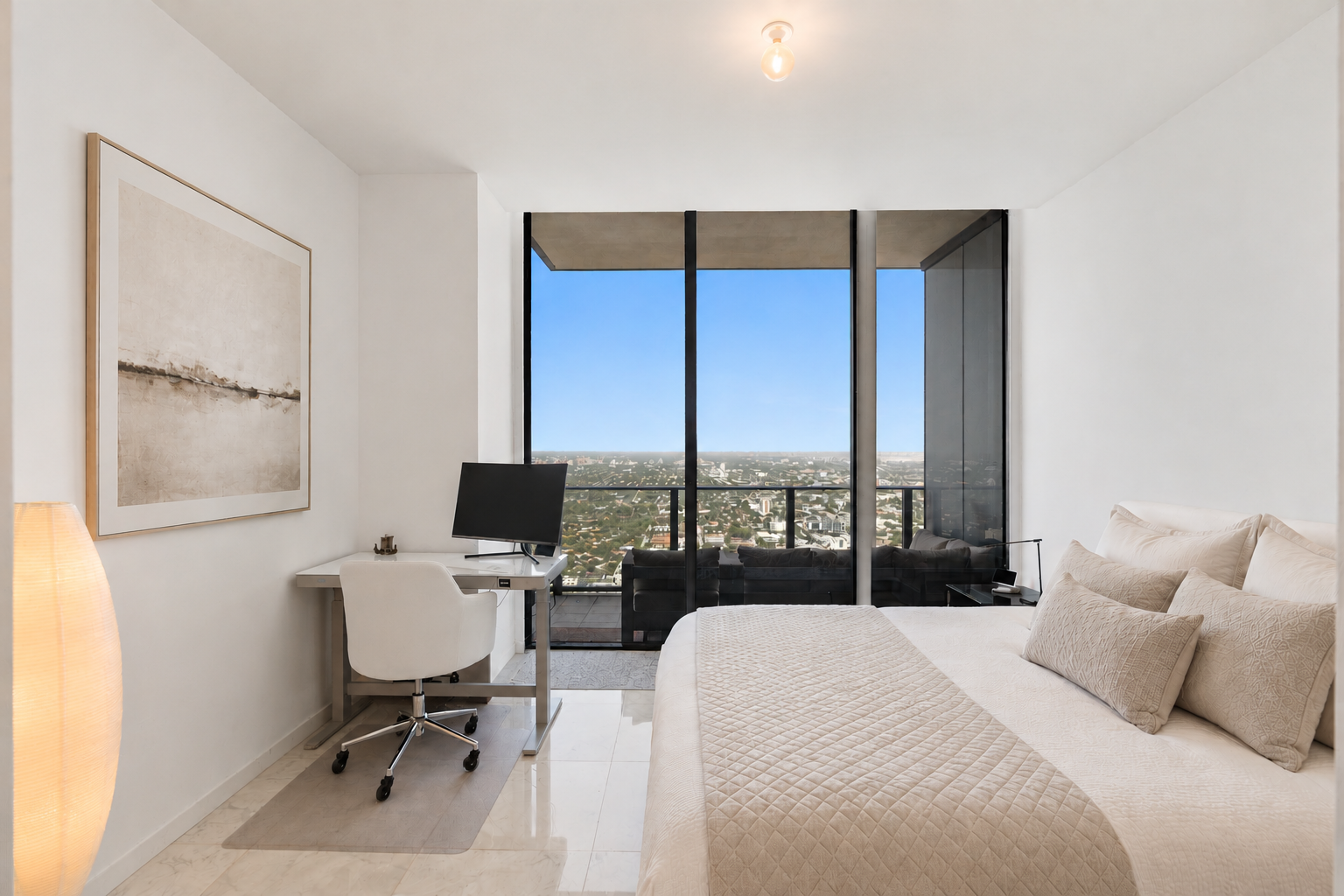 Modern bedroom with large floor-to-ceiling windows showing city view, white bed with beige bedding and pillows, white desk with computer, black sofa on balcony, and a standing floor lamp.