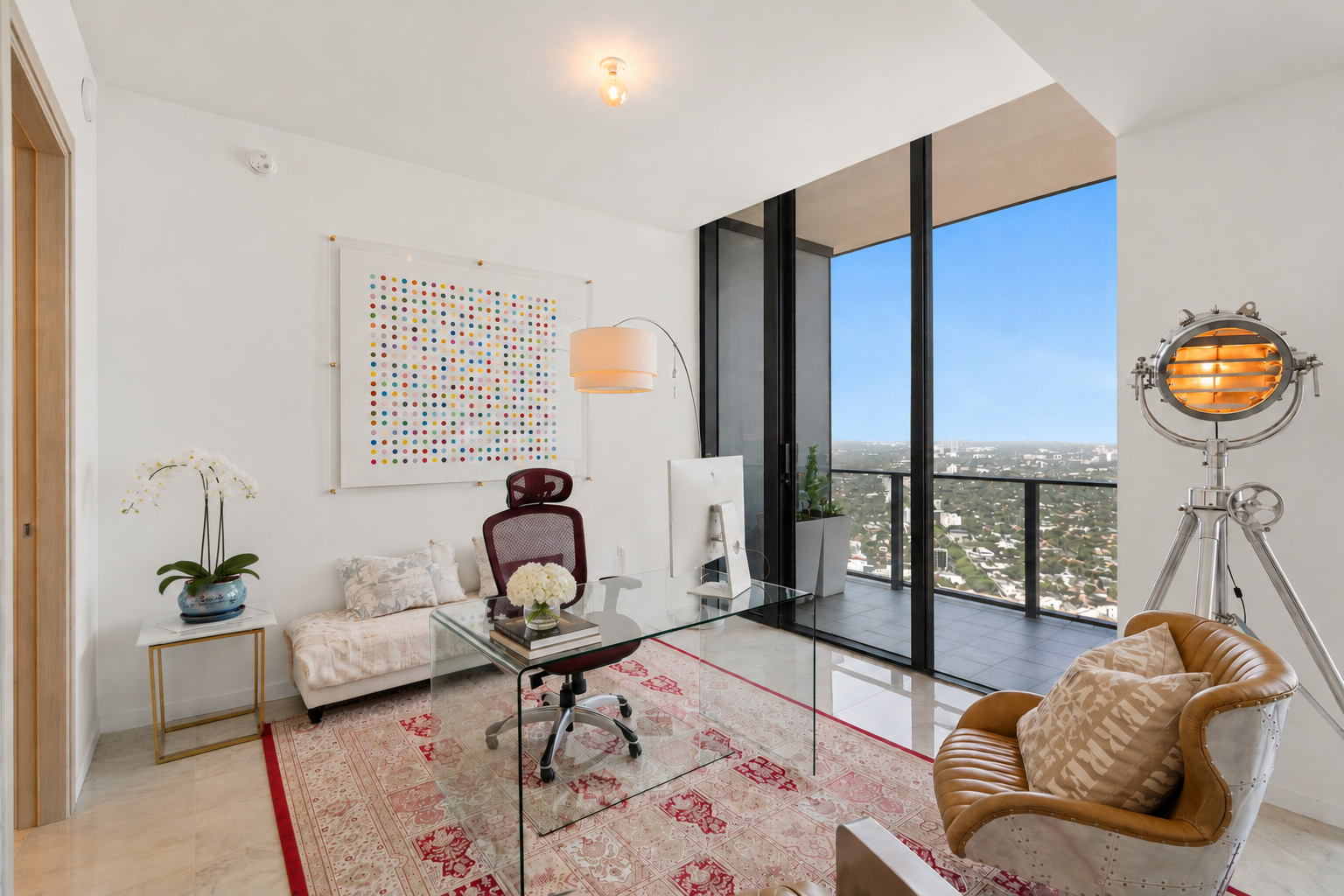 Modern office with glass desk, ergonomic chair, leather armchair, large window with city view, artwork on white wall, potted plant, floor lamp, traditional rug, decorative flower arrangement, and film lighting equipment.