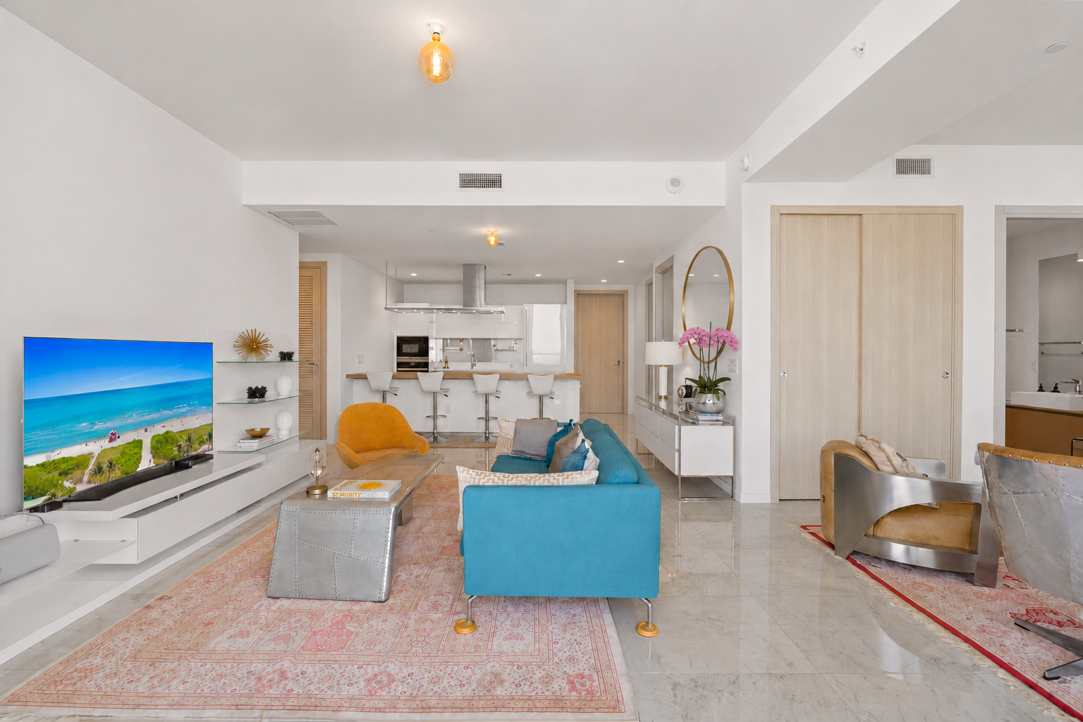 Modern living room with a blue sofa, orange chair, TV displaying beach scene, and bar stools at a kitchen island, decorated with pink flowers and contemporary furniture.