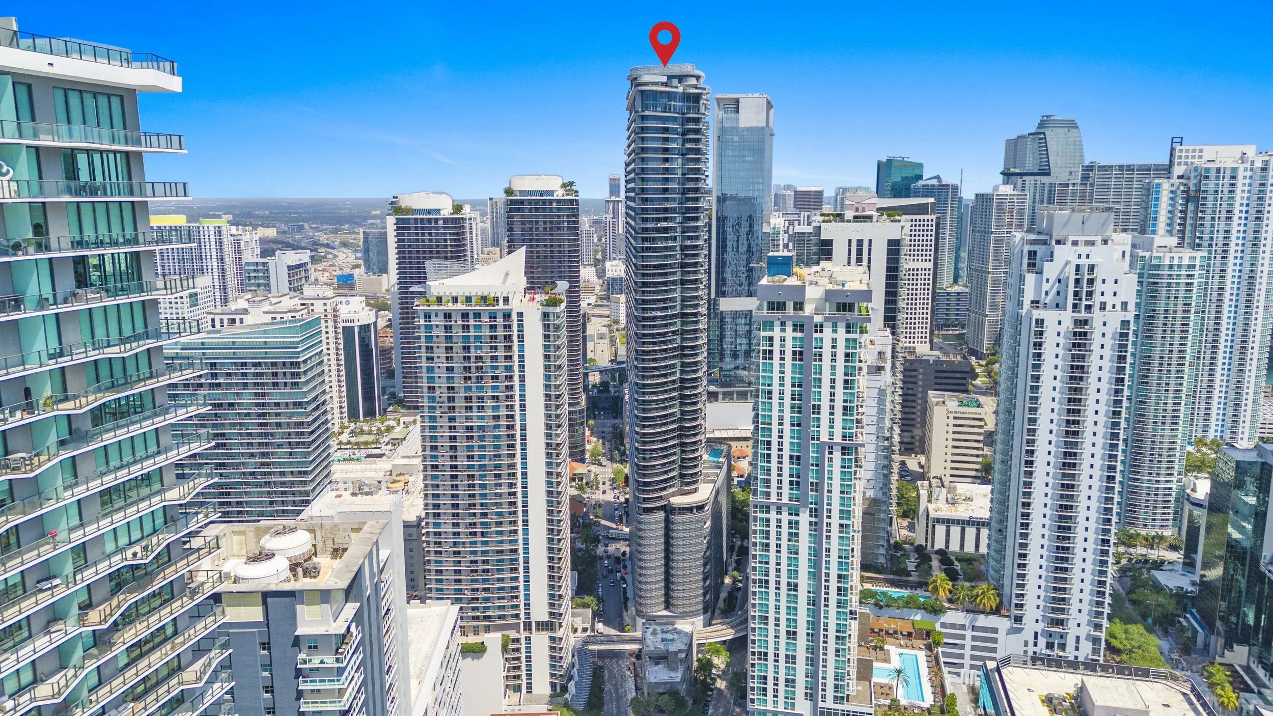 A city skyline featuring numerous high-rise buildings, including a tall building marked with a red location pin on top, under a clear blue sky.