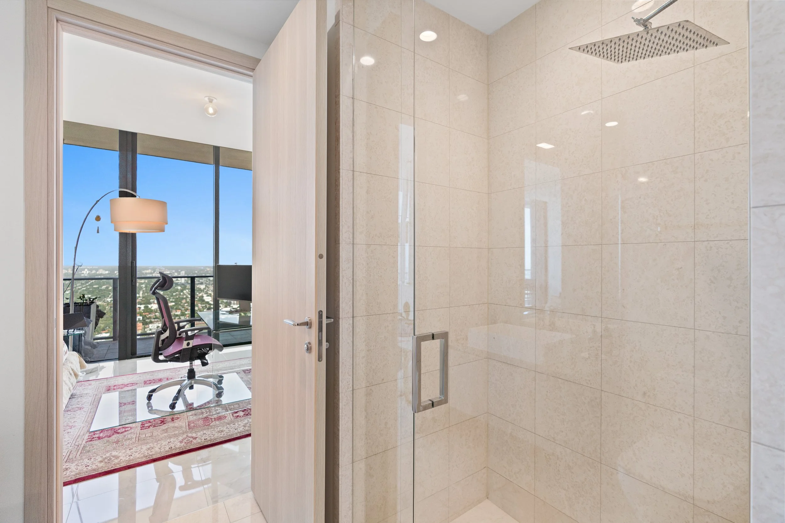 Photo of a modern bathroom with beige tiled shower area, viewed through an open wooden door. The bathroom features a large window revealing a cityscape, and in the adjacent room, a desk with a office chair and floor lamp are visible.