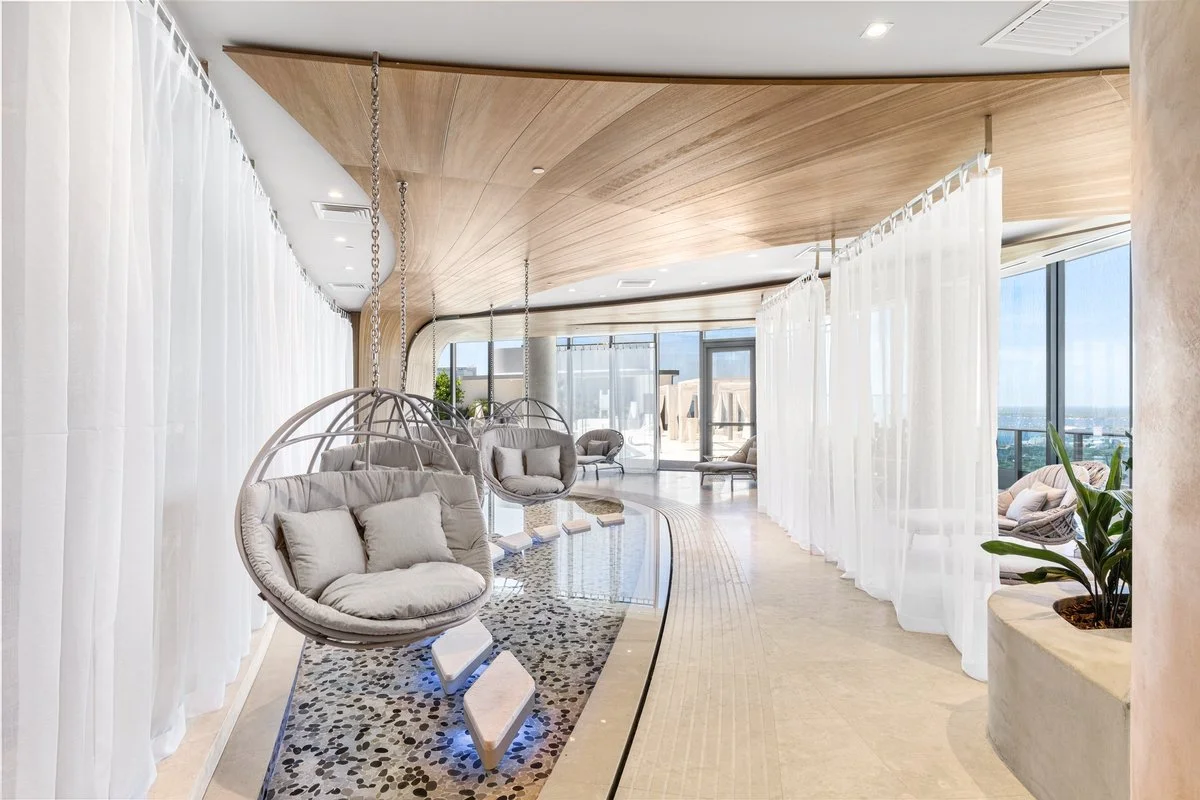 Modern lounge area with hanging cushioned swings, white curtains, and large windows showing a city view.