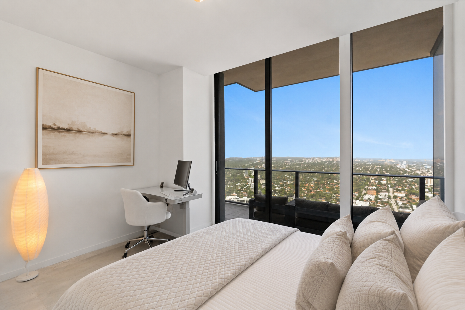 Modern bedroom with a large window overlooking a cityscape, a bed with white linens, a desk with a computer, a chair, and a tall floor lamp.