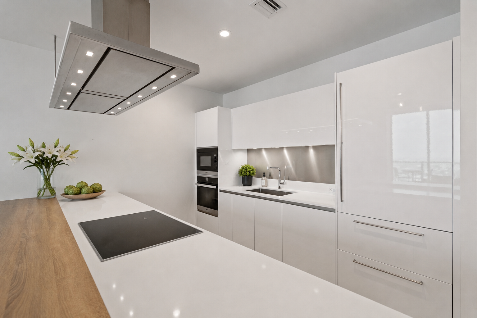 Modern kitchen with white cabinets, a black induction cooktop, built-in oven and microwave, stainless steel backsplash, sink, potted plant, vase with white lilies, and artichokes on a wooden countertop.