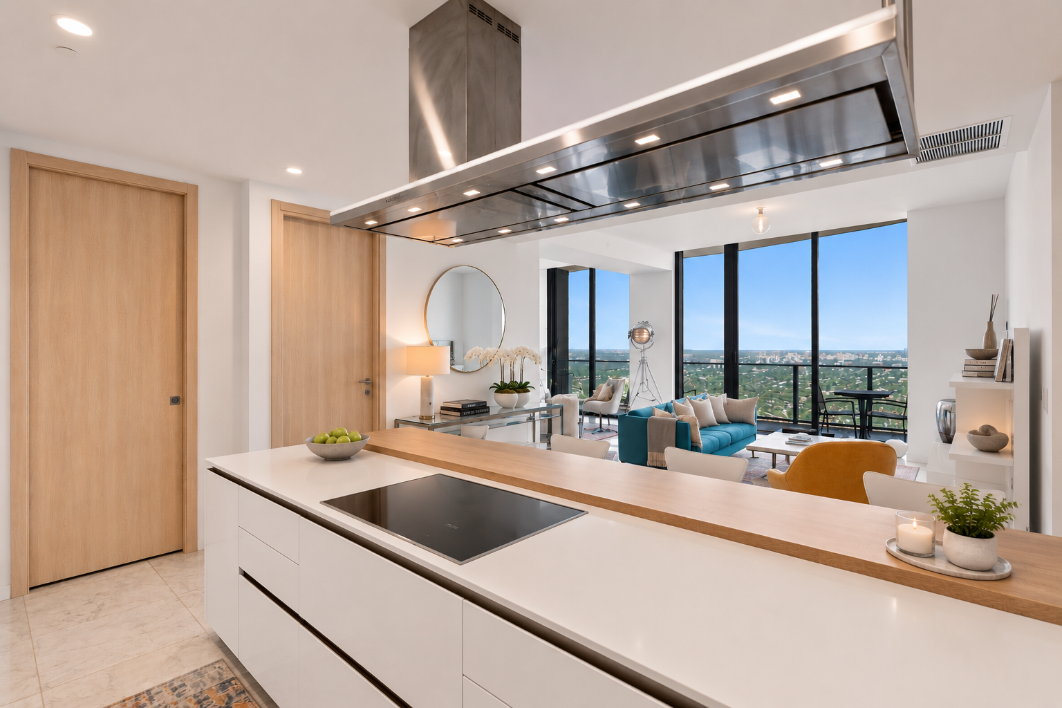 Modern open-concept kitchen and living room with large windows showcasing cityscape views, featuring minimalist decor, a white kitchen island with a black cooktop, a wooden countertop, a bowl of green apples, a round mirror, a table lamp, sofa, armch