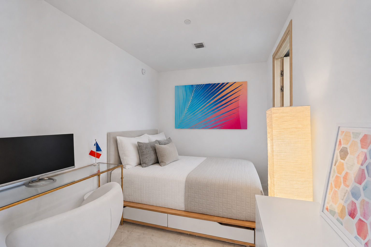 A minimalist hotel room with a white bed, gray and beige pillows, a colorful tropical leaf painting on the wall, a flat-screen TV on a white desk, a small flag, a tall paper lantern, and a framed geometric art piece on a white dresser.