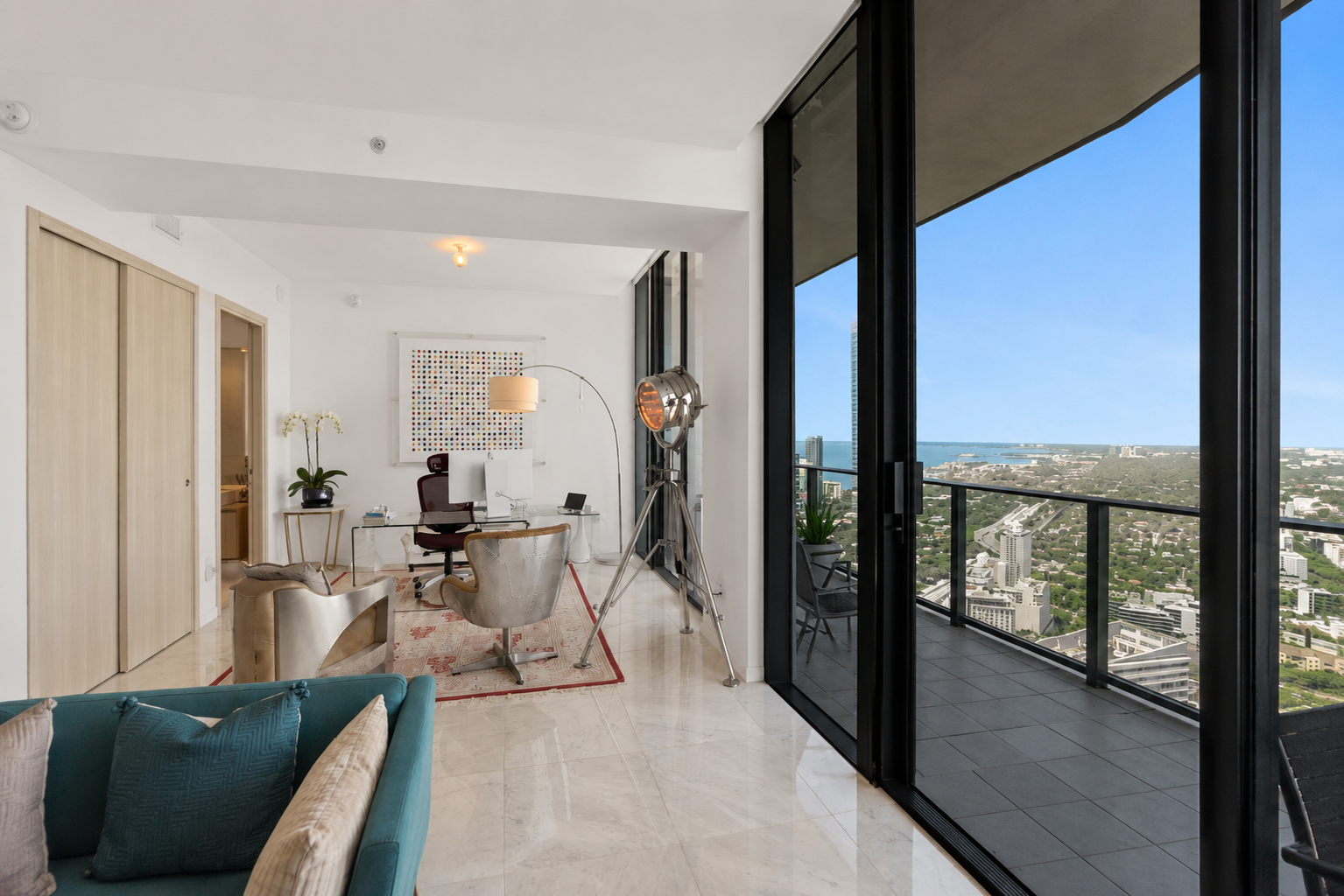 Modern high-rise apartment with a balcony, cityscape view, glass sliding door, office area with a desk, chair, and tall lamp, and a teal sofa with decorative pillows.